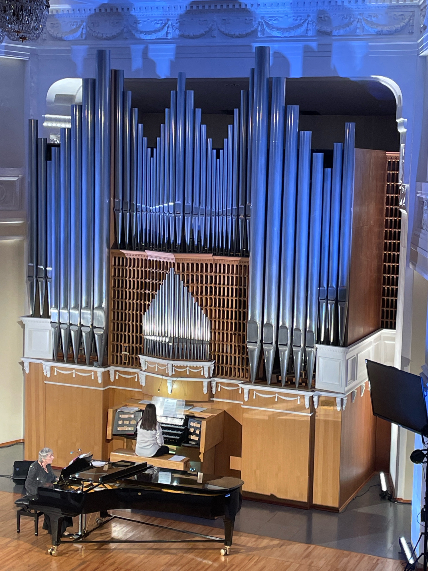 The image shows an interior view of a concert hall where two individuals are performing music. In the foreground, a woman with gray hair is seated at a black grand piano, wearing a dark, sequined top. She is looking towards the piano. To her right and slightly behind her, another woman is seated at an organ console, facing away from the camera. She is wearing a light gray top and is actively playing the organ. The organ itself is a large, imposing instrument with numerous silver pipes of varying lengths and diameters, arranged in sections. The console has multiple keyboards and visible stops. The environment appears to be a formal setting, with ornate architectural details visible on the walls and ceiling, including what looks like a chandelier. The lighting is subdued, with blue and white lights illuminating the organ pipes and architectural features. The floor is a polished wood. In the background, there is a large screen mounted on a stand, and two portable lights are positioned on the floor. The overall scene suggests a musical performance is taking place. The city of Omsk, Russia is not depicted in this image.
