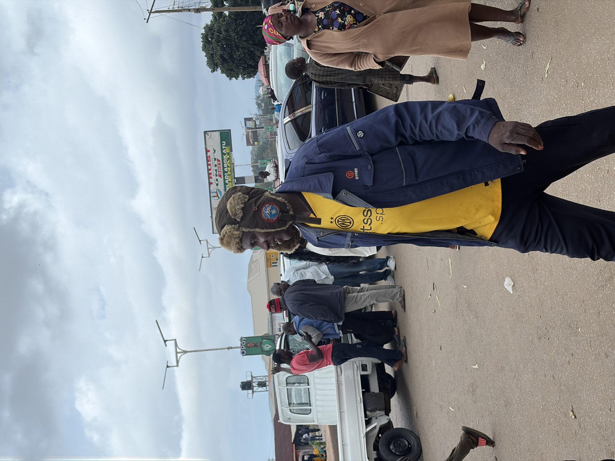 The image depicts an outdoor scene in the city of Gura Top, Nigeria, under an overcast sky. In the foreground, a man in a blue jacket and yellow shirt is walking towards the viewer's right. He is wearing a fur-lined hat and dark pants. To his right, a woman in a tan coat and patterned dress walks away from the viewer. Behind them, a group of people are gathered near a white pickup truck. One man in a red shirt is speaking to another man holding a phone. Several other individuals are standing or walking in the background. The environment includes paved roads, streetlights, a billboard, and various vehicles. The overall impression is a bustling street scene with pedestrian and vehicular activity.