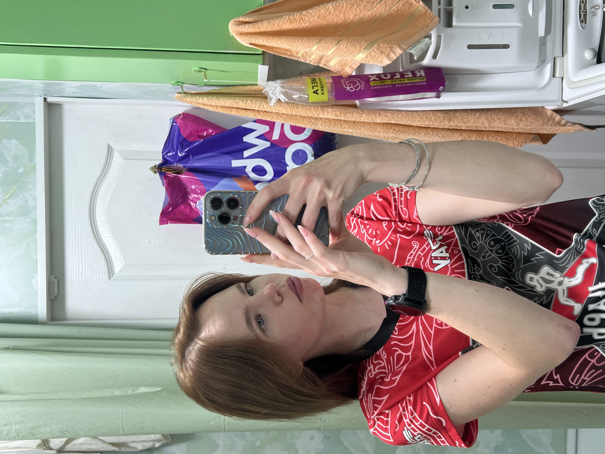 The image shows a young woman taking a mirror selfie in what appears to be a bathroom. She has fair skin, light brown hair, and is wearing a red t-shirt with a black and white graphic design. She is holding a smartphone in her right hand, with her left hand resting on her chest. A watch is visible on her left wrist. In the background, there is a light green shower curtain, a white door, a green cabinet, and two orange towels hanging on hooks. A purple plastic bag with white lettering is also visible hanging on the door. The overall scene suggests a domestic setting, possibly a home bathroom in Chaikovsky, Russia, given the language on some visible packaging which appears to be Russian.