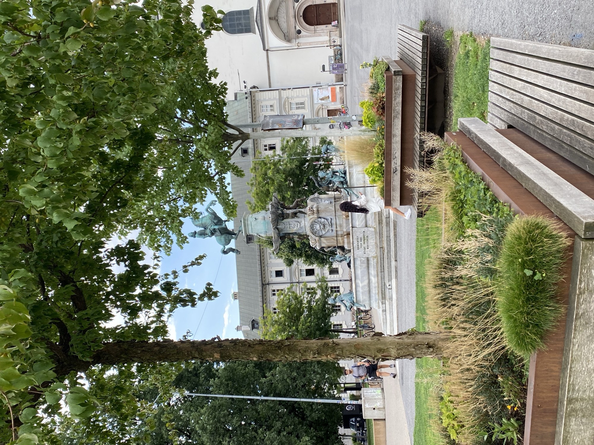 The image captures a view of the fountain in the Innsbrucker Stadtsäule, located in the heart of Innsbruck, Austria. In the foreground, a large metal planter box filled with various grasses and greenery sits on a wooden bench. A tree with textured bark and green leaves partially obstructs the view of the fountain and the surrounding buildings.
In the mid-ground, a bronze equestrian statue stands atop a fountain adorned with figures. The fountain's base is made of marble and features inscriptions. To the right of the fountain, a woman in a white dress is visible, walking away from the camera. Behind the fountain, a light-colored building with multiple windows and a grey roof is prominent. To the far right, a portion of a white church building with an arched wooden door is visible.
In the background, a man in a grey t-shirt and black shorts walks away from the camera, holding a backpack. Bicycles are parked to the left of the fountain. The scene is set in a paved public square with green grass patches. The sky is partly cloudy.