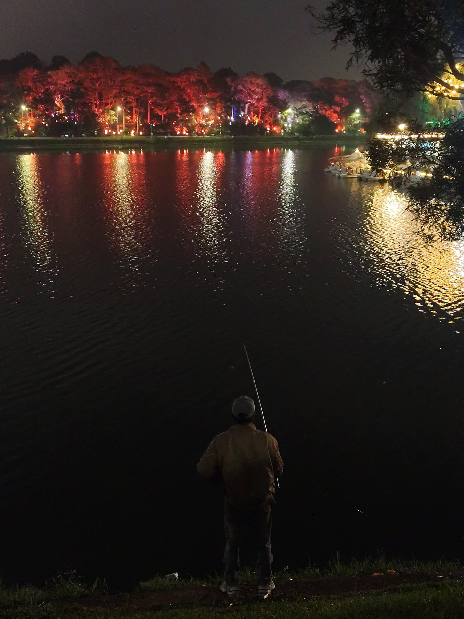 A nighttime scene depicts a lone fisherman standing on the shore of Xuan Huong Lake in Da Lat, Vietnam. The lake reflects the colorful lights illuminating the trees and structures along the opposite bank. The fisherman is wearing a jacket and a cap, facing away from the viewer, holding a fishing rod. The foreground consists of a dark grassy shoreline, while the background is filled with the illuminated lake and distant buildings.