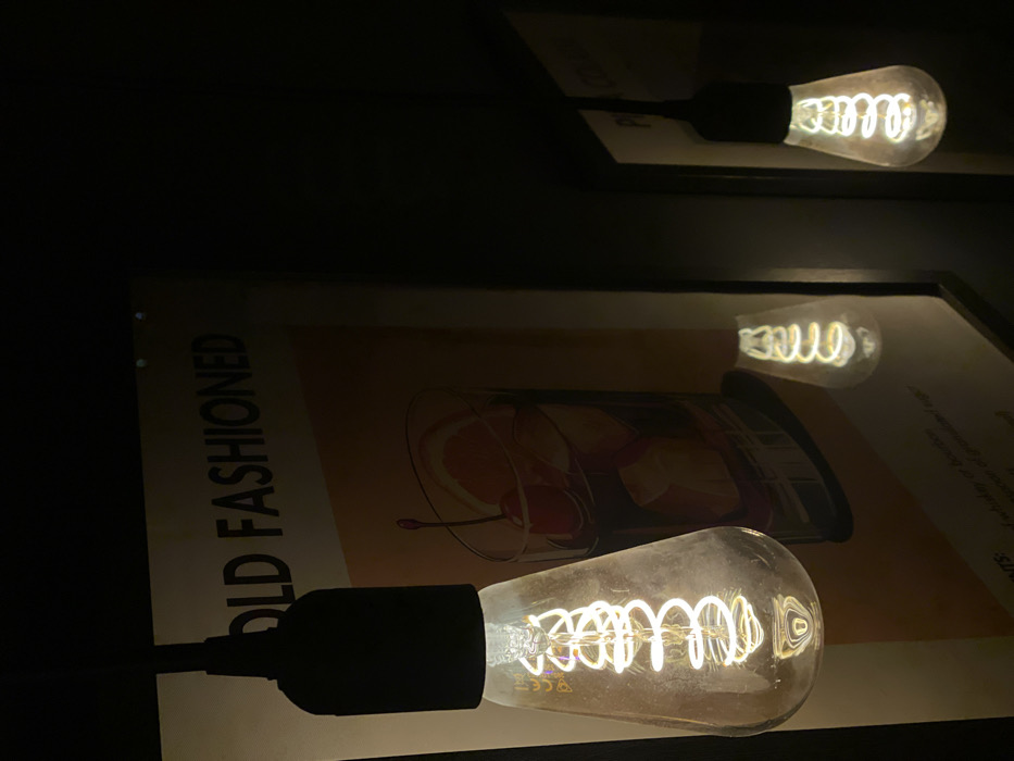 The image depicts a dimly lit interior, characteristic of a bar or restaurant, located in Sofia, Bulgaria. Three hanging, exposed-filament LED bulbs, styled in a vintage Edison design, provide warm illumination. The largest bulb is prominently featured in the foreground, left of center, displaying a luminous spiral filament. Directly behind this bulb is a framed poster illustrating an "Old Fashioned" cocktail, with the words "OLD FASHIONED" clearly visible at the top. The cocktail depiction includes ice, an orange slice, and a cherry. Partial text below the cocktail illustration refers to ingredients such as "whiskey or bourbon." A smaller, illuminated bulb is reflected in the glass surface of this poster. A third, smaller illuminated bulb is visible further to the right in the background. To the far right, a second framed object or poster is partially visible in deep shadow, displaying text that begins with "PI." No individuals, explicit actions, or direct interactions are present in the scene.
