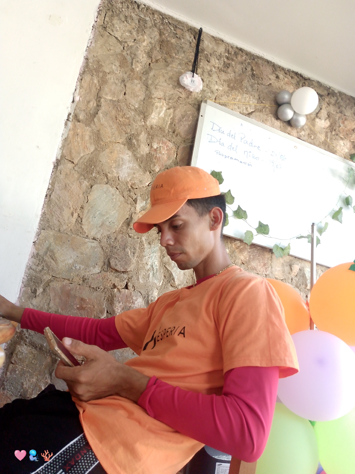 A man wearing an orange t-shirt, long red sleeves, and an orange cap is sitting indoors, looking at his cell phone. The t-shirt has the word "ESPERTA" on it. Behind him is a stone wall with a whiteboard attached. The whiteboard has "Dia del Padre - 21/06" and "Dia del nino - 19/07 Programación" written on it. Balloons in orange, purple, and green are positioned to the right of the man. The location is Playa El Agua, Venezuela.