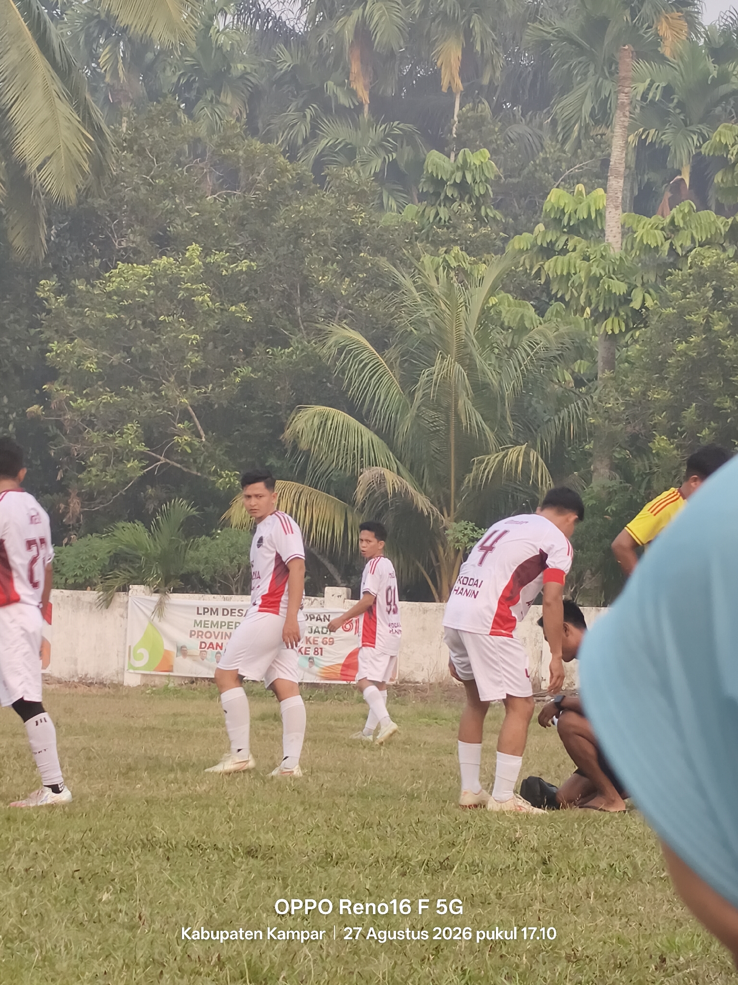 In Penghidupan, Indonesia, a football match is underway on a grassy field. The field is bordered by a white wall with a banner that reads "LPM DESA MEMPER PROVIN DAN" and "OPAN JAD KE 69 KE 81". The players are wearing white and red uniforms, with some sporting the number 4 and 91. In the background, palm trees and other lush vegetation create a dense, tropical backdrop. The sky is overcast, suggesting a humid, possibly rainy day. The photograph captures a moment of intense focus from a player in the foreground, his gaze fixed on the game. Another player, wearing a yellow jersey, can be seen in the distance. The overall scene depicts a local sporting event in a vibrant, natural setting.