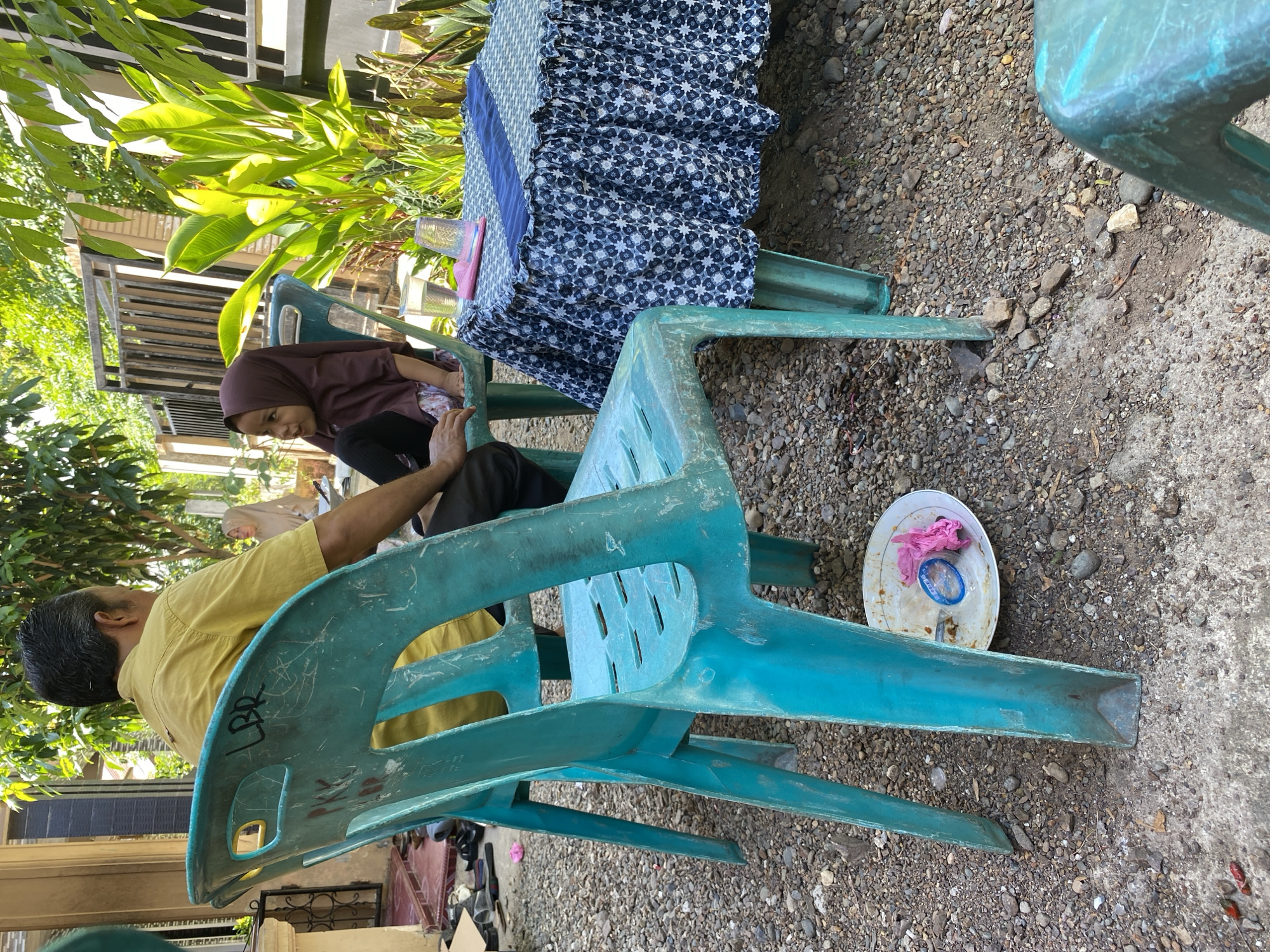 In an outdoor setting, two people are seated on turquoise plastic chairs. An adult male, wearing a yellow collared shirt, is in the foreground, facing away from the camera. A young child, wearing a brown hijab and dark leggings, is seated in a chair to his right, looking to the left. A small, round table covered with a blue and white patterned tablecloth is positioned between them. Two clear plastic cups are on the table, one containing a clear liquid. A pink napkin is also on the table. The ground is covered in gravel and dirt. To the right of the table, lush green plants are visible. In the background, a building with light-colored walls and a dark-colored roof is partially visible, along with a black metal fence. On the ground near the chairs, a white plate contains a pink object and a blue circular item, possibly a condom wrapper. The scene appears to be a casual outdoor gathering or waiting area.