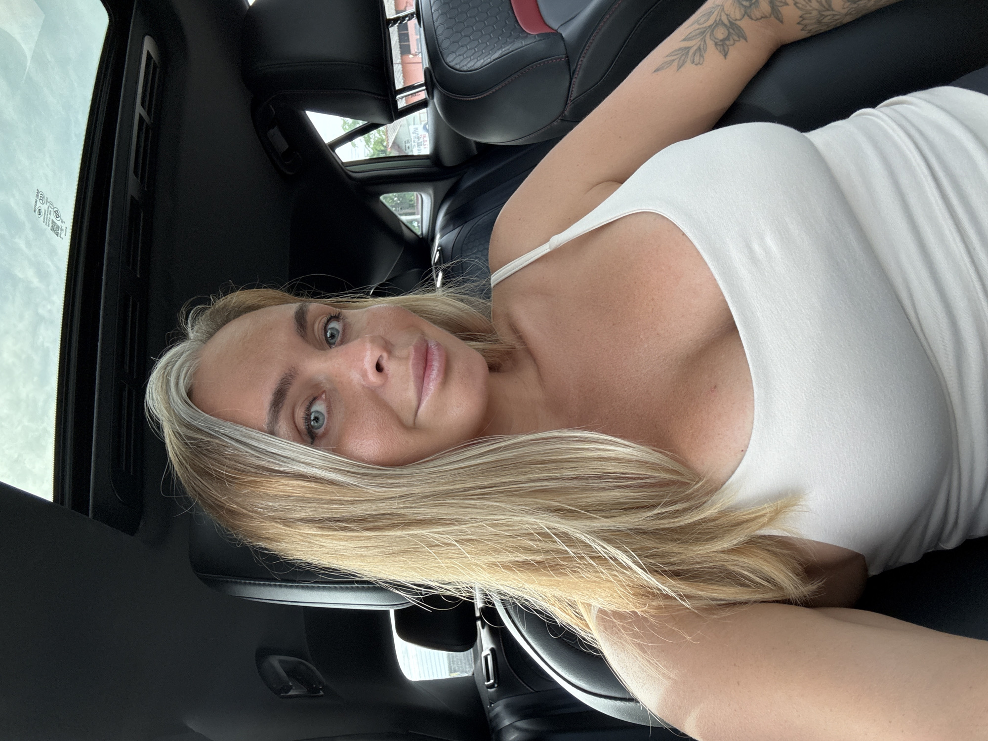 A woman with blonde hair and blue eyes is sitting in the driver's seat of a car, taking a selfie. She is wearing a white tank top, and her right arm, which is tattooed, is visible. The car's interior is dark, with black leather seats and a sunroof. Outside the car, buildings and trees are visible, suggesting an urban or suburban setting in Ko Samui District, Thailand. The sky is cloudy.