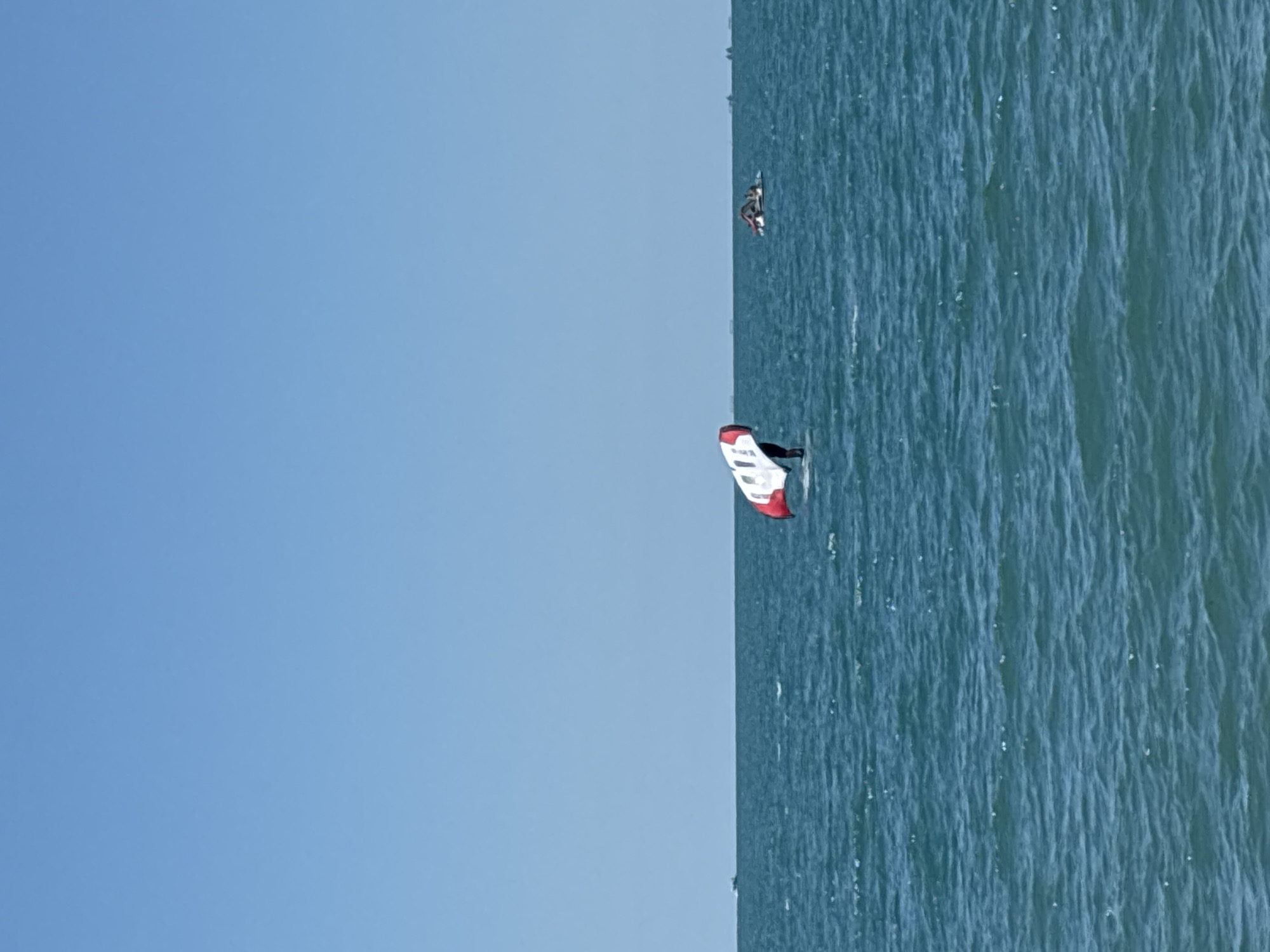 On a clear, sunny day, the vast expanse of the Black Sea at Sochi, Russia, is depicted with gentle waves under a cloudless blue sky. In the middle of the frame, a person is engaged in the watersport of wing foiling, standing on a board with a foil beneath and holding a large wing above their head. The wing is white with red accents. To the right of the wing foiler, another group of people are on a flat, inflatable watercraft, also on the sea. In the far distance, the silhouettes of several ships are visible on the horizon. The overall scene captures a recreational activity on the water.