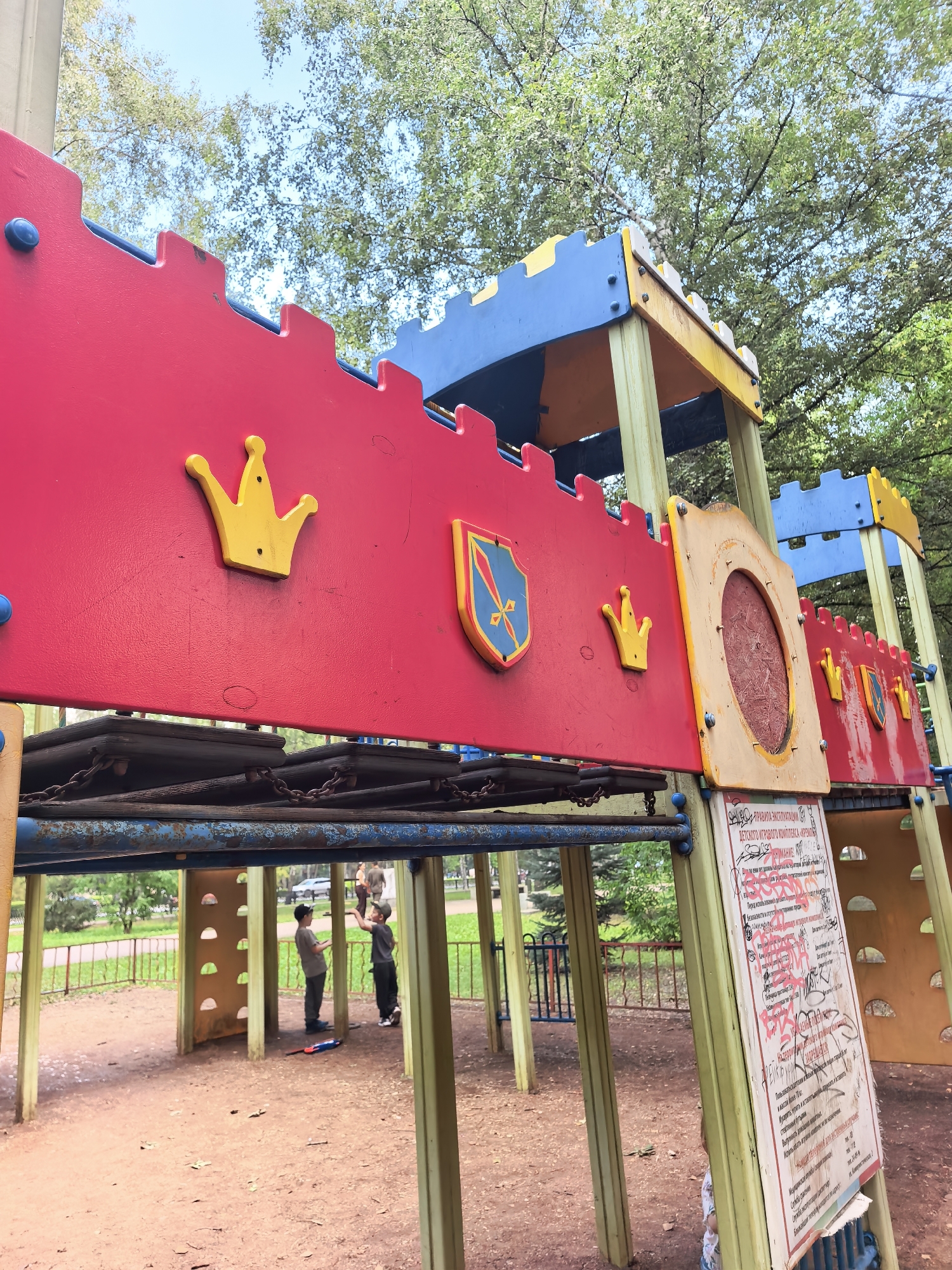 The image shows a colorful playground designed to resemble a castle, situated in an outdoor park. The playground structure is primarily red, blue, and yellow, with decorative yellow crowns and a shield emblem. Beneath the castle facade, metal bars and chains are visible, suggesting climbing or suspension elements. Two young boys are interacting under the structure, one standing with his hands on his hips and the other facing him. A toy rocket lies on the ground nearby. A sign with Russian text, partially covered in graffiti, is attached to one of the support beams of the playground. The background features green trees and a clear sky, indicating a daytime setting. The ground is covered in reddish-brown dirt and scattered leaves. The presence of playground equipment and trees suggests a public park within the city of Sterlitamak, Russia.