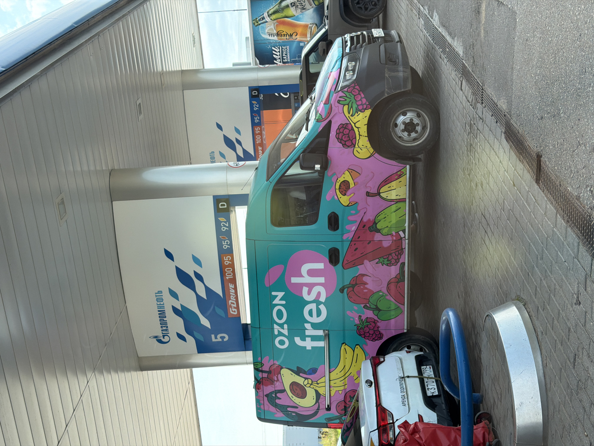 The image shows a turquoise van with a colorful graphic of fruits and berries parked under a white canopy at a Gazpromneft gas station. The van has "OZON fresh" written on its side in large pink lettering. Behind the van, a white Kia car is parked, and a dark SUV is also visible. The gas station pumps are marked with fuel prices. The ground is paved with interlocking grey bricks. The scene is outdoors on a sunny day. The image was taken in Durykino, Russia.