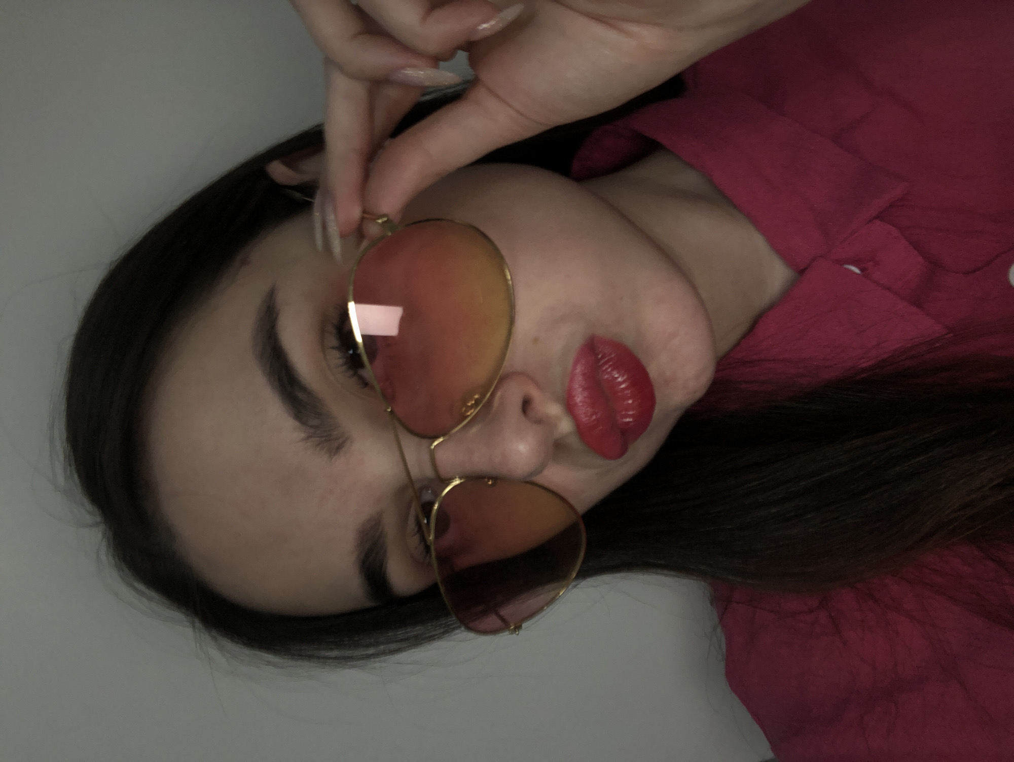A woman is holding a pair of aviator-style sunglasses with gold frames and rose-colored lenses. She has dark hair, pale skin, and is wearing a bright pink shirt. The background appears to be a plain, light-colored wall. There is no visual information in the image to identify the location as Noviy, Russia, or any other specific place.