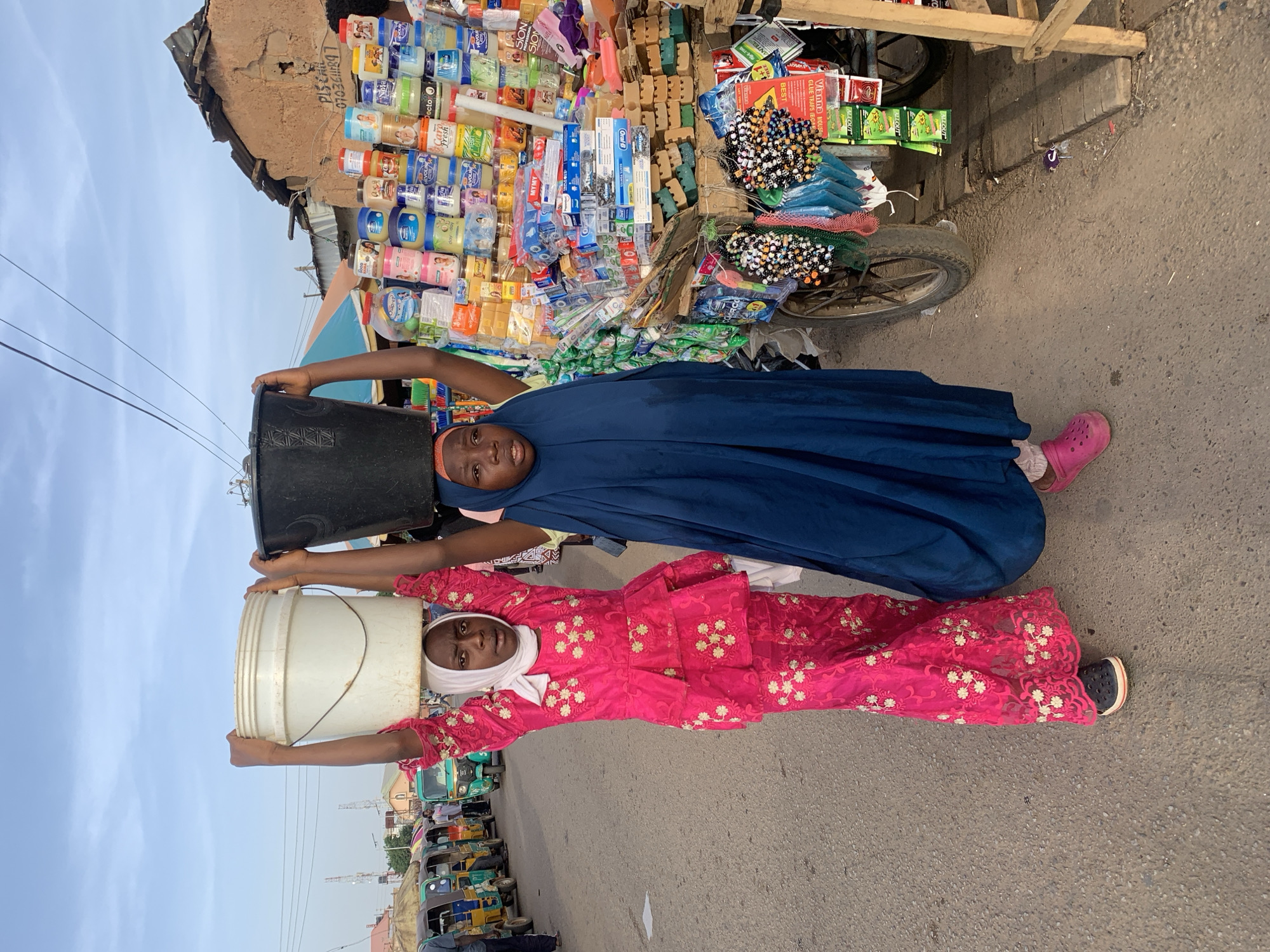 Two girls are carrying buckets on their heads in Gura Top, Nigeria. The first girl, in the foreground, wears a pink dress with white floral detailing and a white headscarf, and she carries a white bucket. The second girl, behind the first, wears a blue garment and carries a black bucket. Both girls are facing the camera. Behind them is a cart full of consumer goods for sale. In the background, there is a row of auto rickshaws.