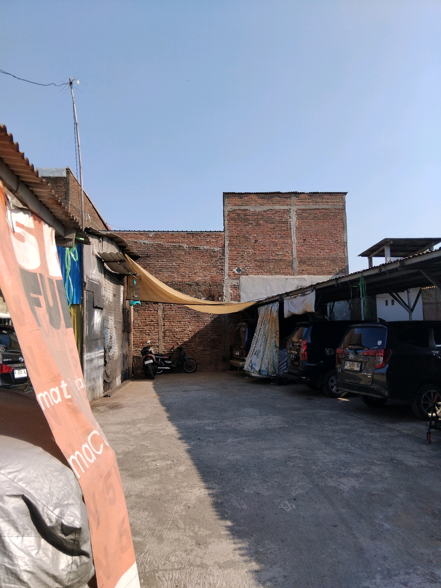 The image depicts an outdoor scene in what appears to be a semi-urban or industrial area, possibly a workshop or a yard, in Kepuh Kiriman, Indonesia. The sky is clear and blue. In the foreground, a large orange banner with white text is partially visible on the left side, and a silver-gray tarp covers an object. The ground is paved with concrete, showing signs of wear and tear.
To the left, a corrugated metal building stands, with some blue and green materials hanging near its entrance. A black vehicle is parked in front of this building. Further into the scene, a light-colored awning or tarp is stretched between buildings, casting a shadow. Beyond this, a brick building forms the background.
In the middle ground, several vehicles are present. A dark-colored motorcycle is parked near the brick building. To the right, two dark-colored cars are parked under a covered structure with a corrugated roof. One of these cars is a Toyota Avanza with license plate number N 1698 VG. Another dark vehicle is partially visible on the far right. A bird cage hangs from the awning above the cars.
The overall impression is one of a functional, somewhat weathered outdoor space.