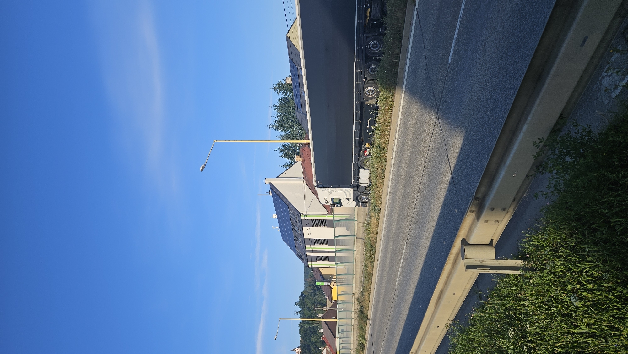 A daytime, eye-level, vertical, full shot captures a semi-trailer truck traveling on a paved road under a clear blue sky. The truck, primarily white with a black trailer, is in the right half of the frame, moving from left to right. In the background, several buildings are visible, some with solar panels on their roofs. A yellow lamppost stands to the right of the truck. The foreground shows a grassy embankment with a metal guardrail. The scene appears to be on the outskirts of Prešov, Slovakia, likely along a highway or major road given the presence of the guardrail and the semi-trailer truck.