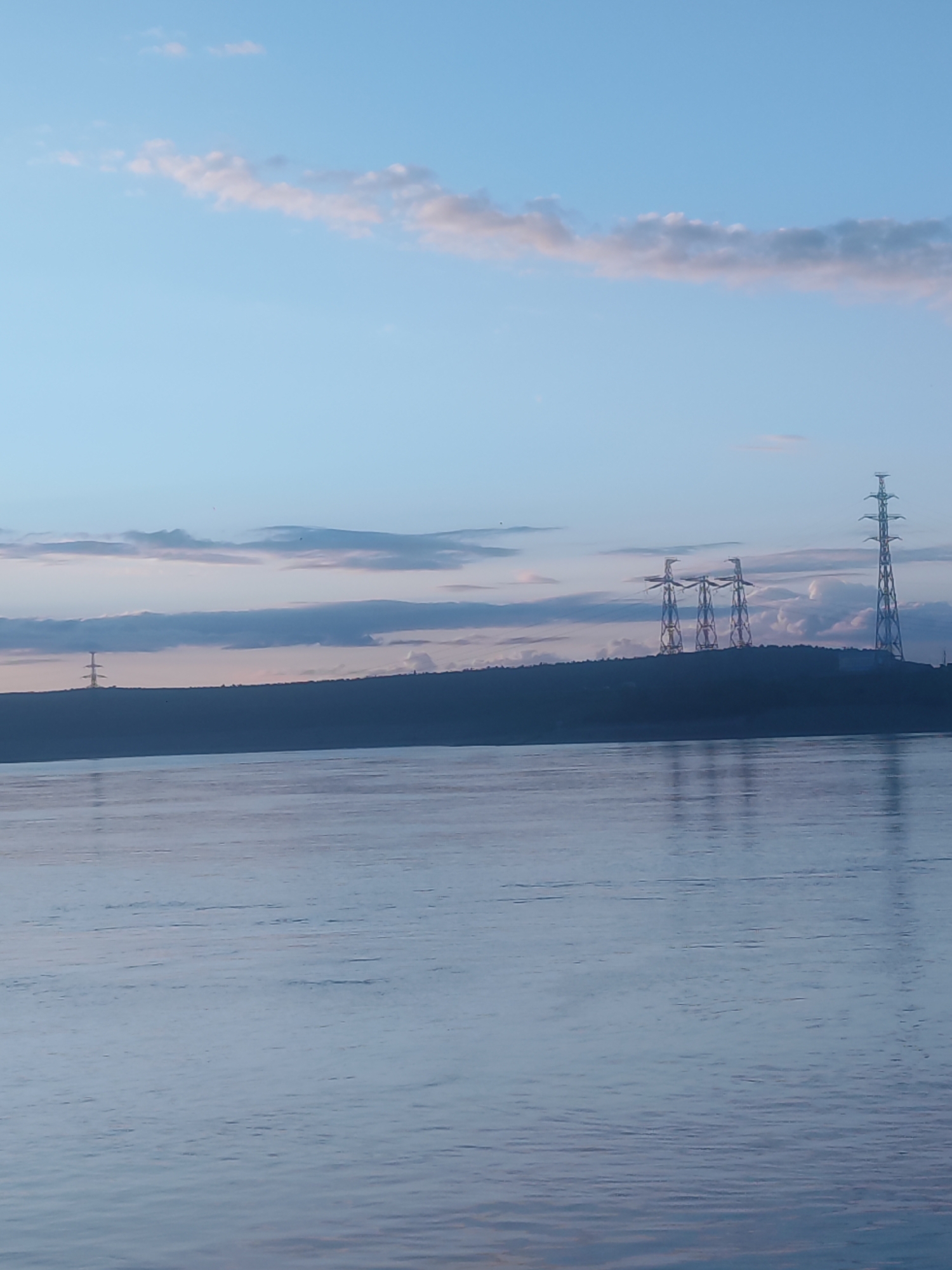 The image presents a tranquil, wide landscape featuring a body of water, a distant landmass, and several power transmission towers under a vast sky, likely at dawn or dusk in Noviy, Russia.
The foreground is dominated by a calm, expansive body of water, possibly a river or a lake. Its surface is a soft bluish-grey, with gentle ripples and faint reflections of the sky and structures above.
Across the middle of the frame, a dark, silhouetted strip of land, appearing as a forested hill or shoreline, stretches horizontally. Positioned on this distant land are several tall, intricate power transmission towers. To the far left, a single multi-tiered pylon stands. Towards the mid-right, a group of three distinct, modern-looking power towers with multiple legs is visible. Further right, an exceptionally tall, slender tower rises against the horizon. These towers, despite being man-made, are integrated into the vast natural scene.
The sky, filling the upper half of the image, is a soft, clear light blue. Scattered across it are elongated, wispy clouds that exhibit subtle hints of pink and grey, characteristic of the ambient light during either sunrise or sunset. The overall lighting is diffused and gentle, contributing to a serene and somewhat contemplative mood.
There are no people or specific activities taking place. The scene emphasizes the vastness of nature alongside human infrastructure. No text is visible in the image.