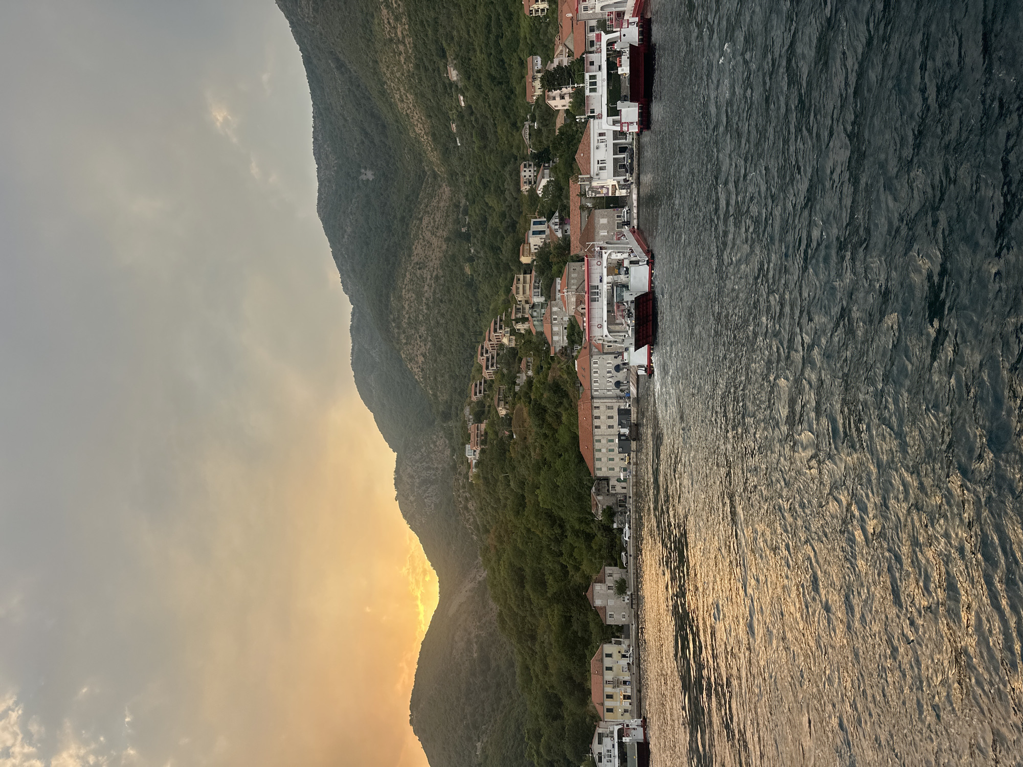 The image depicts a waterfront scene in Jošice, Montenegro, during a sunset or sunrise. The sky is filled with dramatic clouds, with a warm orange glow emanating from the sun, which is partially obscured by the mountains. A range of lush green mountains, dotted with houses, forms the backdrop. In the foreground, the dark, rippling water reflects the sky's colors. Along the shoreline, a row of buildings with red-tiled roofs lines the water's edge. Several ferries are docked or approaching the shore. The overall impression is one of a tranquil coastal town bathed in the soft, warm light of dawn or dusk.