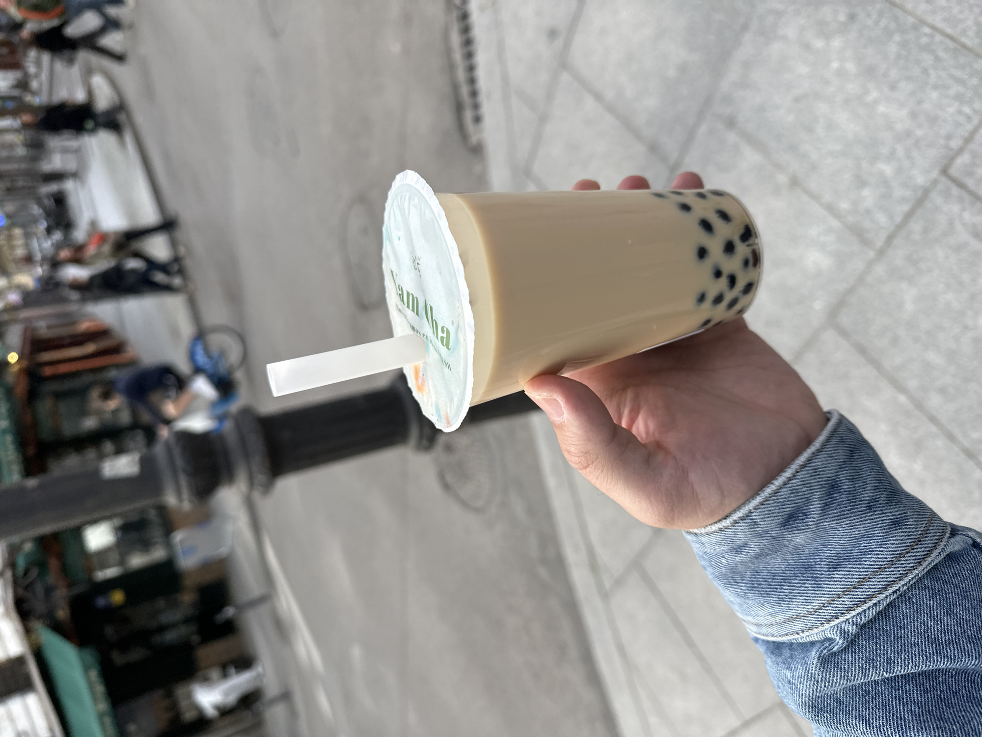 An individual wearing a denim jacket holds a cup of bubble tea. The location appears to be an urban street, possibly in Madrid, Spain. A light post and other people can be seen in the background. The bubble tea is in a clear plastic cup with a sealed lid and a white straw.