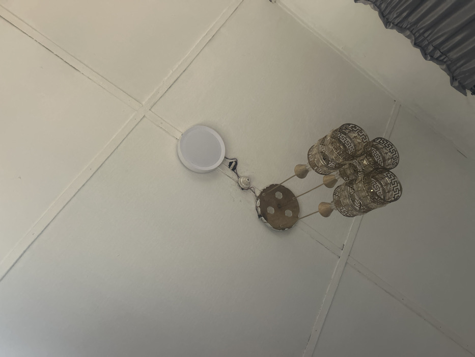 This image shows a white ceiling in an interior space located in Ilishan, Nigeria. The ceiling structure features a visible grid pattern from panels or tiles. A multi-pendant chandelier is suspended, consisting of a round, metallic base from which three cylindrical light shades hang. Each shade is gold-toned with a repeating Greek key-like geometric pattern. Adjacent to the chandelier, a white, circular, flush-mounted fixture, appearing as either a recessed light or a ceiling speaker, is installed. Exposed electrical wiring and an uncovered electrical junction are visible between the chandelier's base and the circular fixture, indicative of an exposed or unfinished electrical installation.