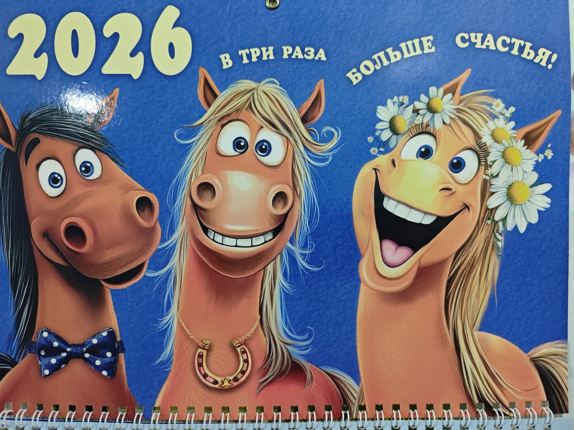 The image is a close-up of a calendar page featuring three cartoon horses on a bright blue background. The leftmost horse, a reddish-brown color, wears a blue and white polka dot bowtie. The middle horse, a lighter shade of brown, has blonde hair and a gold horseshoe necklace. The rightmost horse, also a lighter brown, has blonde hair adorned with a daisy crown. Text in yellow letters across the top reads "2026" and "В ТРИ РАЗА БОЛЬШЕ СЧАСТЬЯ!". The bottom edge of the calendar shows a silver spiral binding and a portion of the next page's white border. The image is well-lit, with no discernible environmental context outside of the calendar page itself, and therefore, no information about its location in Polyarny, Russia can be inferred.