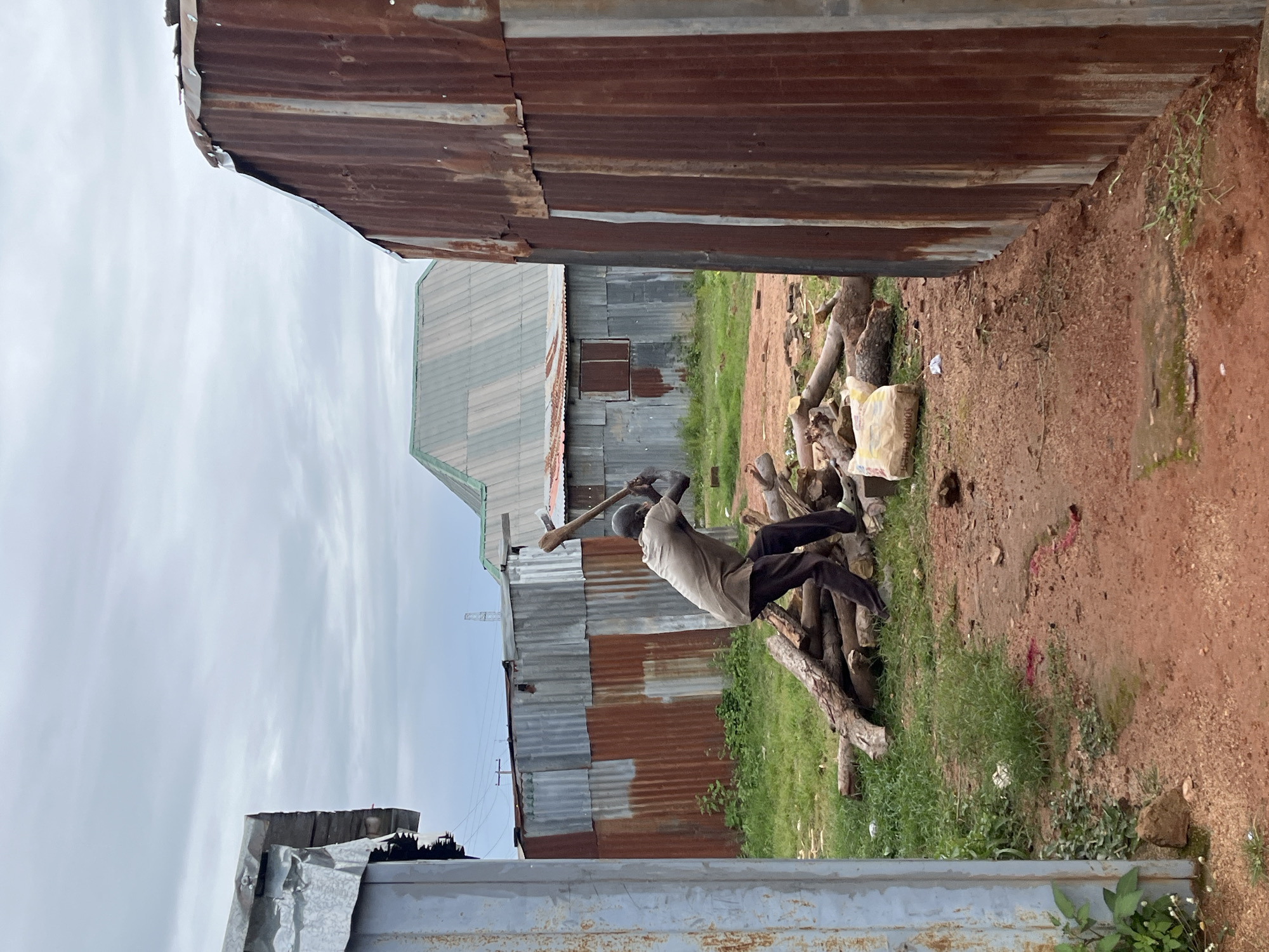 In Barakin Akawo, Nigeria, a person is chopping wood with an axe. The scene is set amongst corrugated metal structures and scattered logs. The ground is a mix of red earth and green grass, with some discarded items present, including a bag of cement. The sky is overcast.