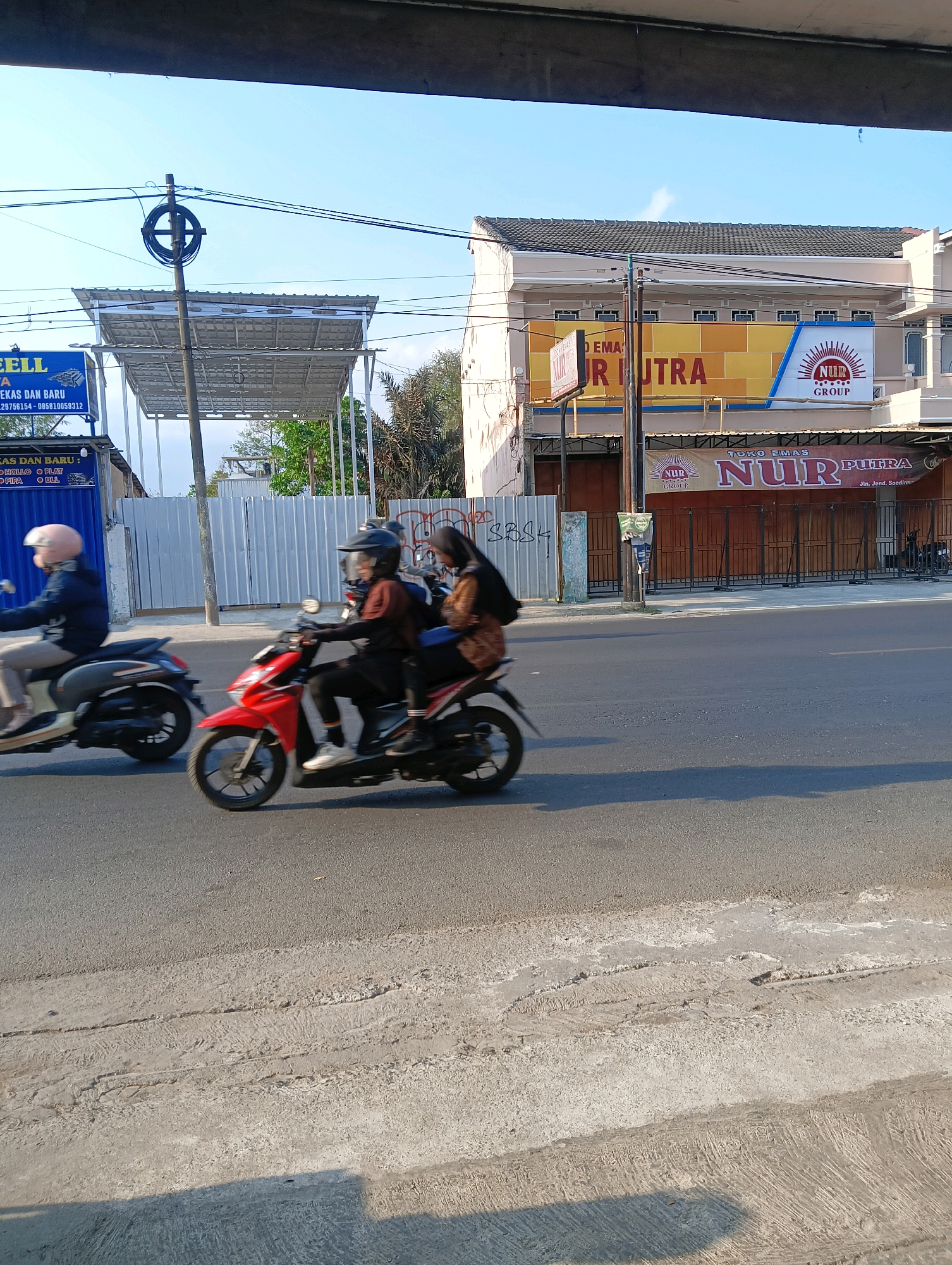 In Sokaraja Kidul, Indonesia, two motorcycles are traveling on a paved road. The first motorcycle, a red scooter, is carrying two individuals. The rider is wearing a dark helmet and a brown long-sleeved shirt, while the passenger in the back is wearing a dark headscarf and a brown jacket. The second motorcycle, a gray scooter, is being ridden by a single person wearing a pink helmet and a dark jacket. Behind the motorcycles, several commercial buildings are visible. One building features a large yellow banner with the text "NUR PUTRA" and a red and white logo. Another building has a smaller banner with the same logo and text, along with "TOKO EMAS NUR PUTRA" and an address. The sky is clear and blue. The road surface shows signs of wear and tear, with some cracked sections. Overhead, electrical wires are strung across the scene.