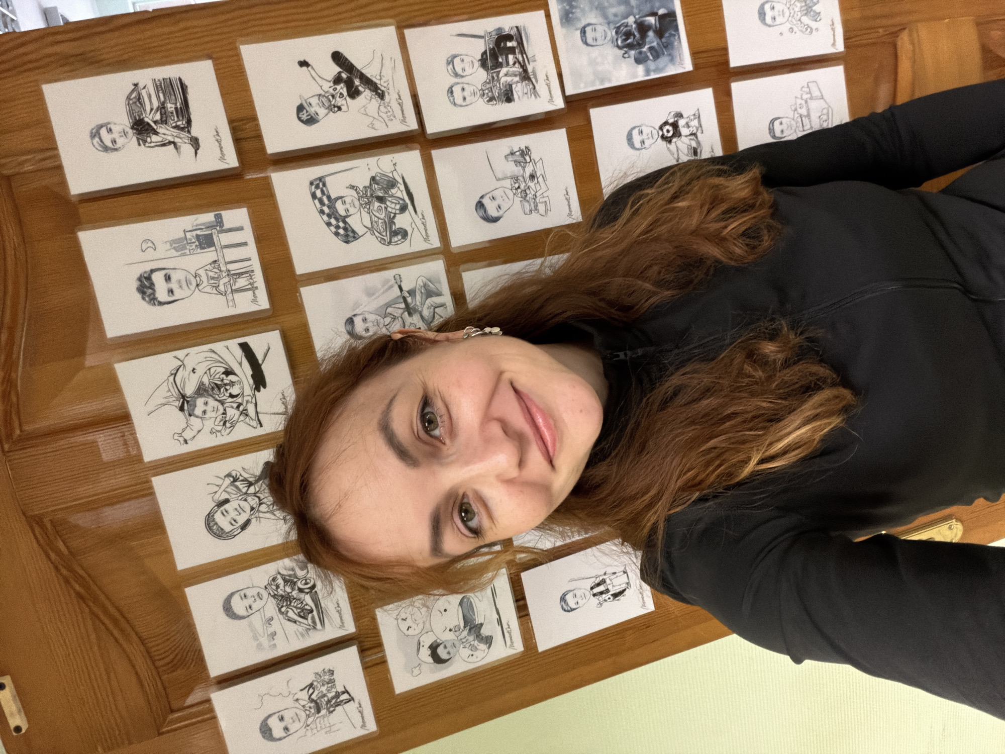 A woman with long, reddish-brown hair is posing for a selfie. She is wearing a black long-sleeved shirt. Her face is close to the camera, showing her eyes, nose, and mouth. Behind her is a wooden surface (possibly a door) covered with approximately 18 small, white cards with black and white illustrations on them. The illustrations seem to depict caricatures of a person engaged in various activities like driving, skating, playing musical instruments, and playing sports. The location is Chaikovsky, Russia. There is a light green wall visible in the lower right corner of the frame. The cards are arranged in a grid-like pattern.