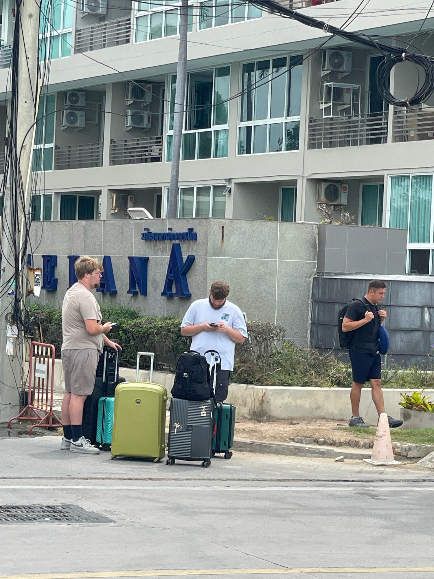 This media file depicts three individuals standing near a building with a prominent sign. The building appears to be a hotel or apartment complex, judging by the multiple balconies and air conditioning units visible on its facade. The sign in blue lettering, which reads "CLEAN K" and some Thai text above it, is mounted on a stone-like textured wall.
Two men are in the foreground, each with their mobile phones in hand. The man on the left, wearing a light brown t-shirt and shorts, is standing beside several pieces of luggage: a black suitcase, a teal suitcase, and a bright green suitcase. The man in the middle, wearing a white t-shirt and dark shorts, is also looking at his phone and has a black backpack resting on the ground beside him, along with a black and grey suitcase. To the right, a third man, dressed in a black t-shirt and dark shorts, is walking away from the camera, carrying a black backpack and a blue bag. He is also looking down, presumably at his phone.
The scene is set outdoors, with a paved area in front of the building and some greenery and landscaping behind the luggage. A red metal barrier is visible to the left of the frame, and a light orange traffic cone is placed on the right side of the pavement. The lighting suggests it is daytime, possibly overcast or late afternoon, as there are no harsh shadows. The presence of multiple pieces of luggage indicates that the individuals may be arriving at or departing from the location, possibly travelers. The overall impression is one of people waiting or preparing for transport. The location context is Pattaya City, Thailand.