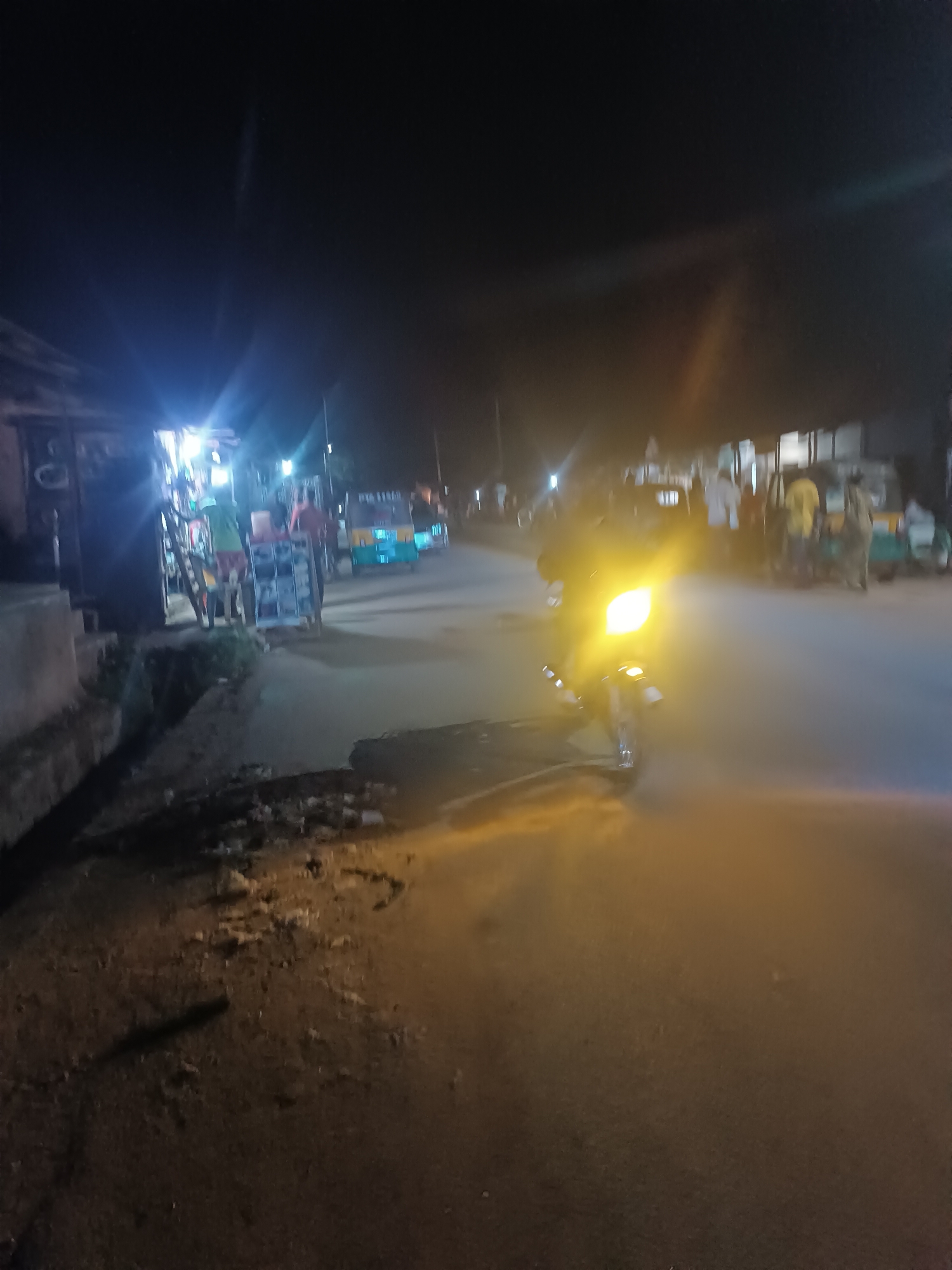 The image depicts a night scene in Barikin Gidar Kanat, Nigeria. A motorcycle with a bright headlight is in the foreground. Street vendors with mobile carts are lit by bright lights on the left side of the street. Tuk-tuks and people are visible further down the street on both sides. The road surface appears to be paved. The sky is dark.