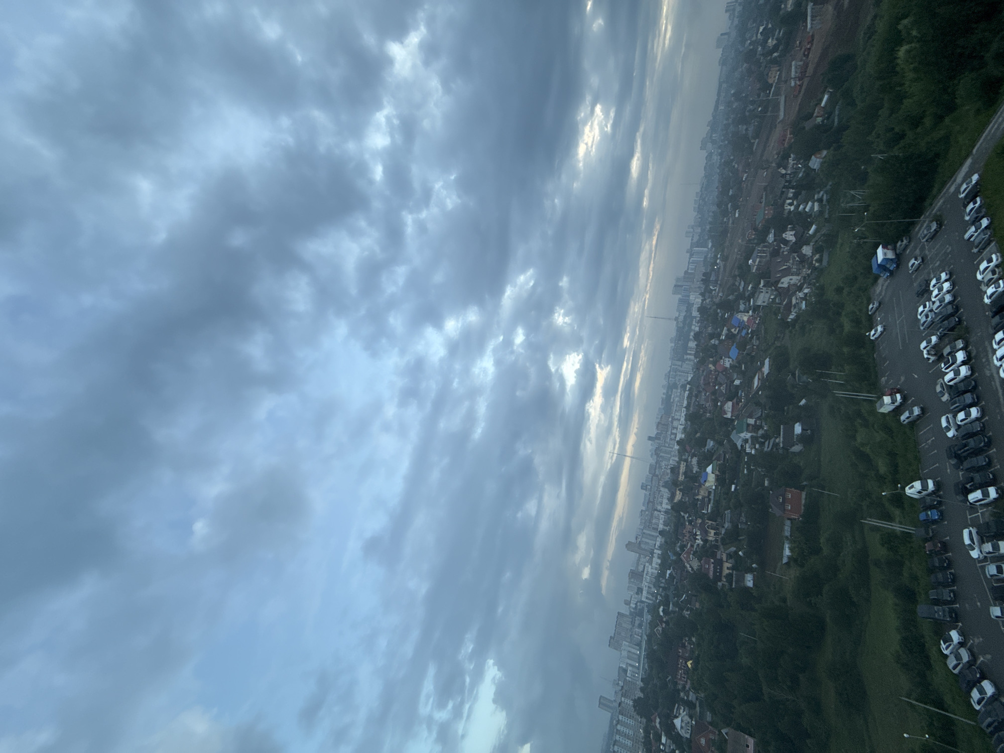 This is a high-angle, landscape orientation photograph taken in Perm, Russia, showing a city skyline and a parking lot under a cloudy sky. The time of day appears to be early morning or late afternoon, as the sun is partially visible through breaks in the dense clouds, casting a soft light. The weather is overcast, contributing to a subdued atmosphere.
The scene is divided into two main areas: the sky and the ground. The sky occupies the left and upper portion of the image, filled with heavy, gray clouds. Shafts of light penetrate the clouds in the upper right portion, suggesting the sun is low on the horizon.
The ground area, on the right and bottom of the image, reveals a sprawling urban and semi-urban landscape. In the foreground, a large parking lot is filled with numerous cars, mostly white and black, parked in rows. Some of the cars are visible in the cropped images, showing their typical shapes and sizes. Beyond the parking lot, the city unfolds with a mix of residential buildings, some appearing to be houses with red roofs and others taller apartment buildings in the distance. Green trees and vegetation are interspersed throughout the city, particularly along the edges of the built-up areas. Construction sites, with visible machinery and building materials, are also present, indicating ongoing development. The overall impression is of a large, lived-in city with a mix of residential, commercial, and natural elements. There is no visible text in the image.