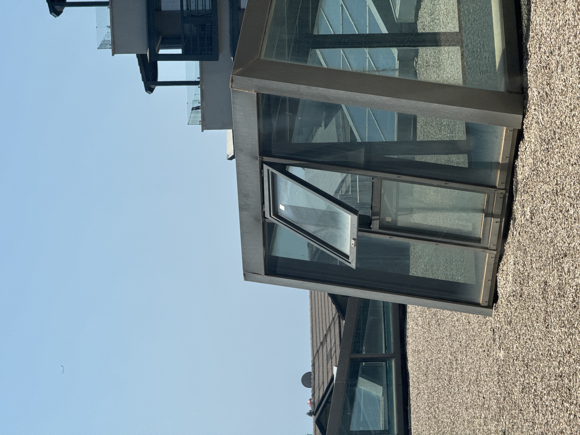 A group of modern glass skylights with metal framing are installed on a gravel-covered rooftop. In the foreground, one skylight is partially open, revealing a glimpse of the interior. In the background, a multi-story building with balconies and glass railings rises against a clear blue sky. A faint airplane is visible in the upper left quadrant of the sky. The scene is set in the city of Aksu, Turkey.