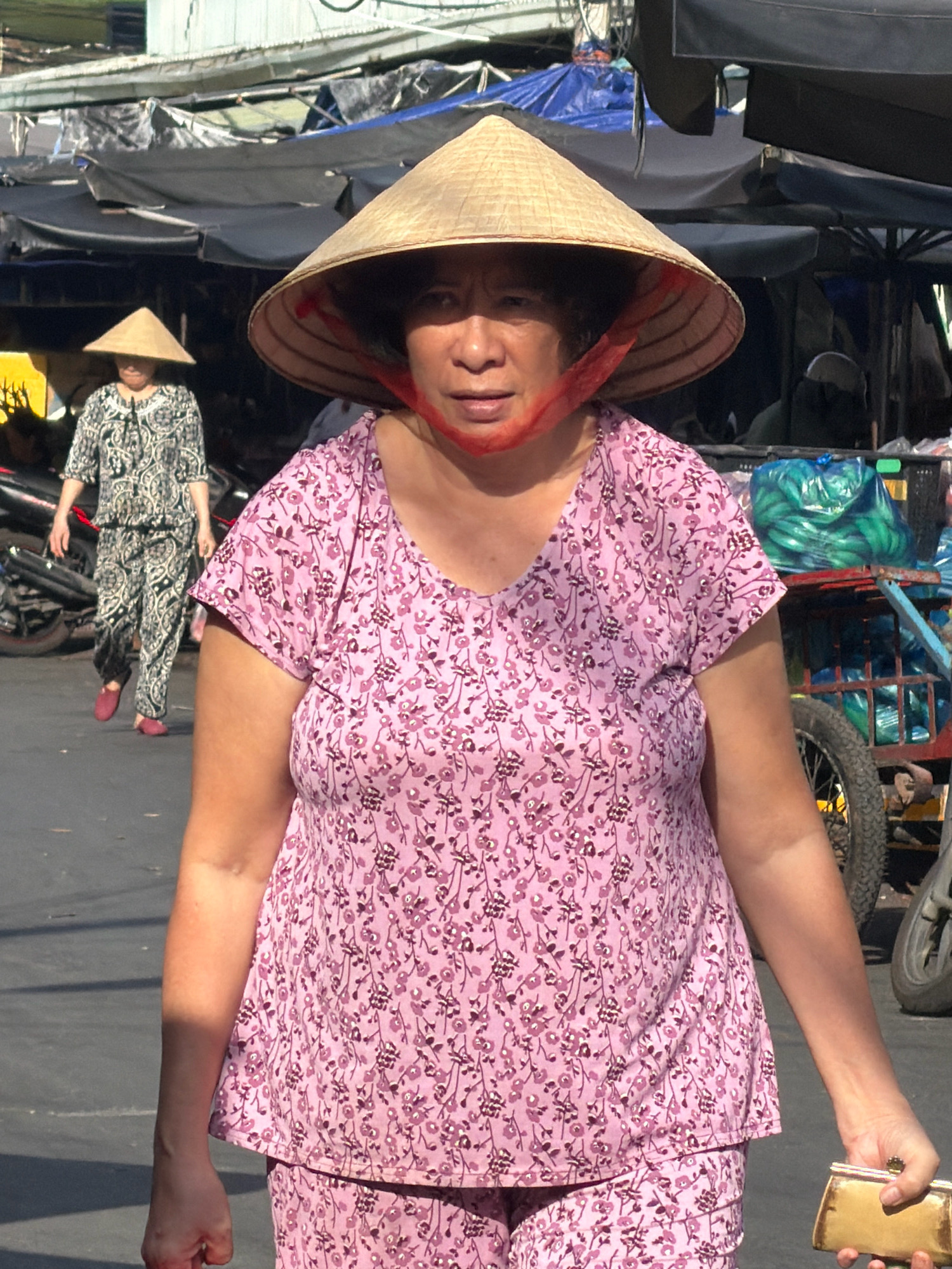 In a sunlit outdoor market in Nha Trang, Vietnam, a woman wearing a conical hat and a pink floral-patterned outfit is the central subject. Her hat, made of woven straw, is secured with a red string around her chin. She is holding a small, golden clutch purse in her right hand. Her left arm is bent, with her hand resting on her hip. To her left, another woman, also wearing a conical hat and a black and white patterned outfit, walks away from the camera. Behind the foreground subject, a blue cart filled with green produce is visible, along with the wheel of a motorcycle and more market stalls. The ground is paved with asphalt. The scene appears to be a busy, daytime market environment.