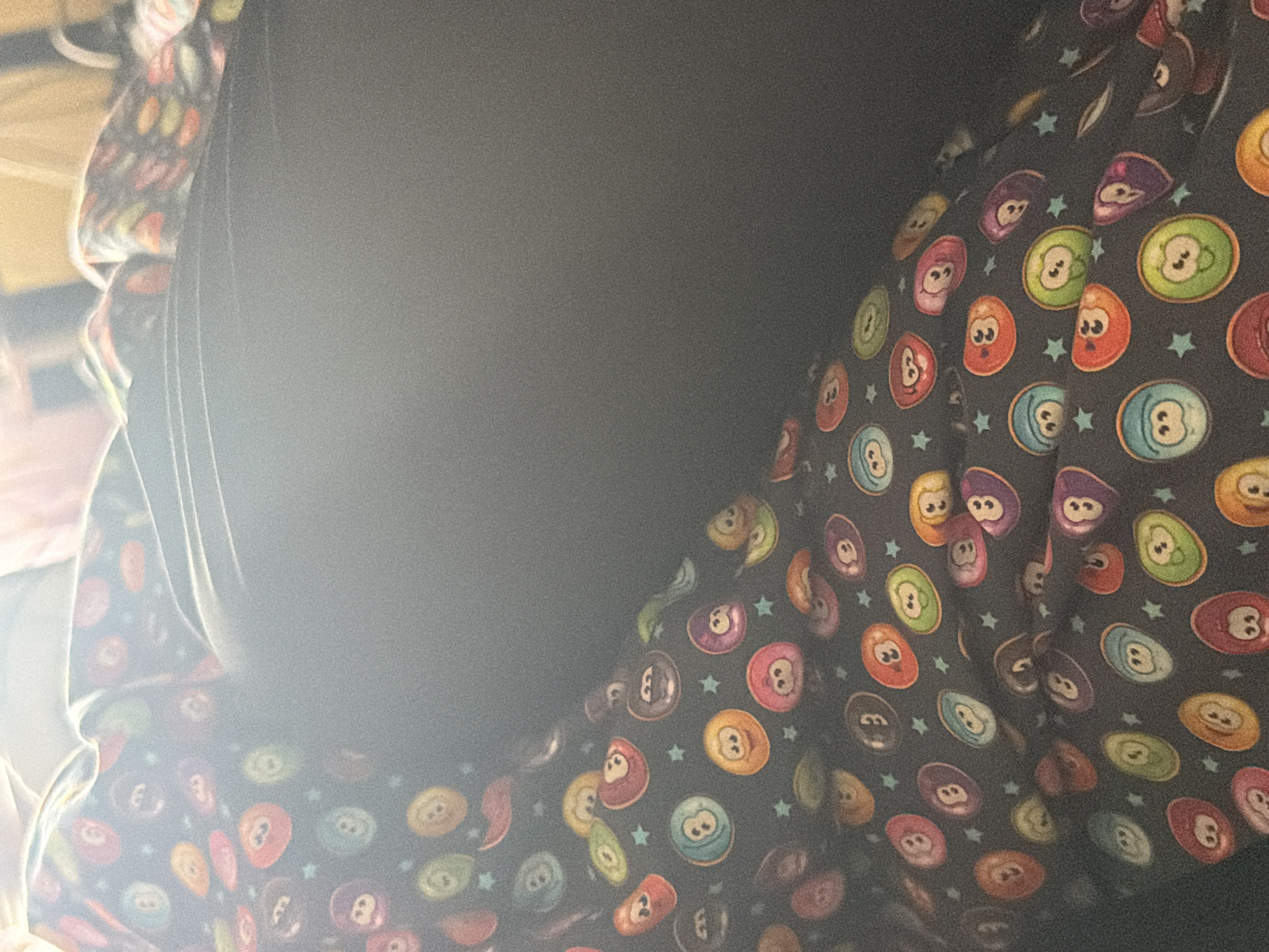The image displays a close-up of two fabric items in an indoor setting. A large portion of the frame, particularly the left and center, is occupied by a plain, dark fabric, likely black or dark grey, which appears to be a piece of clothing or a blanket. This dark fabric exhibits some texture and is significantly affected by a bright light source on the left, causing strong glare and overexposure that obscures some details.
To the right, and partially visible on the left, is another fabric item with a distinct pattern. This fabric has a dark background, likely black or a very dark grey. It is covered with a repeating pattern of colorful, cartoon-like billiard balls. These balls come in various colors such as red, orange, yellow, green, light blue, purple, and pink. Each billiard ball prominently features a number, primarily "8" but also "2" and possibly others, and is depicted with a simple, expressive face, adding a whimsical element. Small, light blue star shapes are interspersed between the patterned billiard balls.
The arrangement of the fabrics suggests they are draped over an object, creating folds and contours. No specific activity or event is taking place; the image is a static capture of these textiles. The strong illumination from the left suggests the picture was taken during the daytime. No specific location cues are visible in the image beyond an general indoor environment.