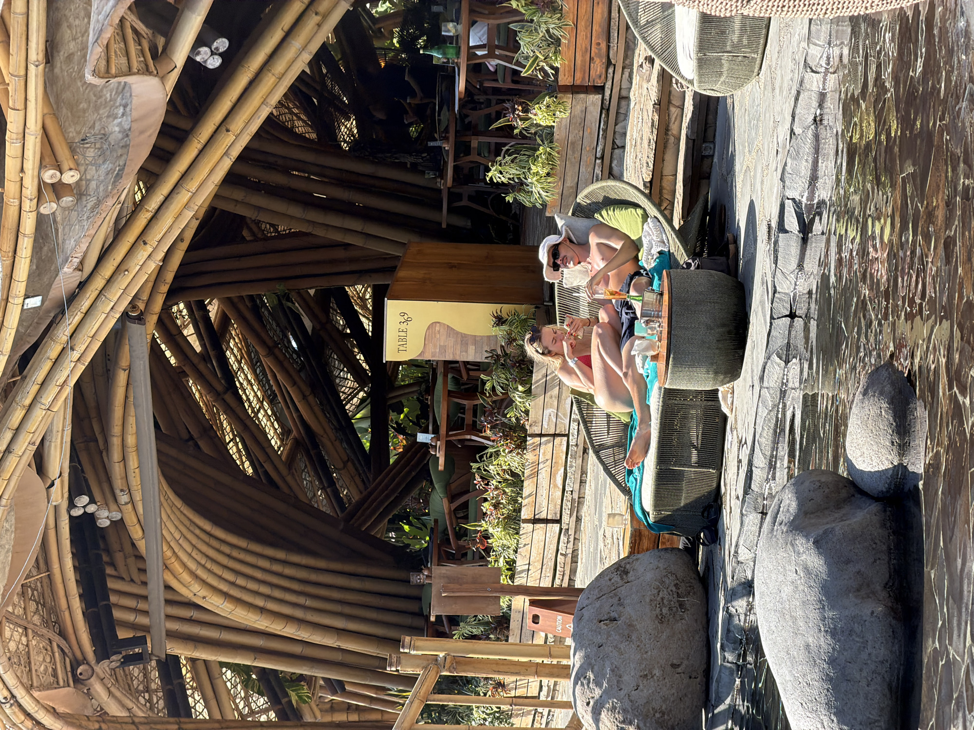 The image depicts an outdoor seating area, likely a restaurant or cafe, with a distinctive bamboo-constructed roof. In the foreground, a shallow body of water with large rocks is visible.
Two individuals are seated on a lounge chair to the right of the frame. The person on the left, a female, is wearing a red bikini and is looking down at her phone. The person on the right, a male, is wearing a white hat and sunglasses, and appears to be holding a drink. They are positioned at a circular table with drinks and an ice bucket.
In the background, there are several tables and chairs set up, suggesting a dining or seating area. The architecture prominently features woven bamboo elements, creating a natural and organic aesthetic. Lush green plants are also visible throughout the space, adding to the tropical atmosphere. A sign with "TABLE 369" is mounted on a wooden structure. The ground is paved with stone. The lighting suggests it is daytime with sunlight casting shadows.
The location is identified as the city of Kediri, Indonesia.