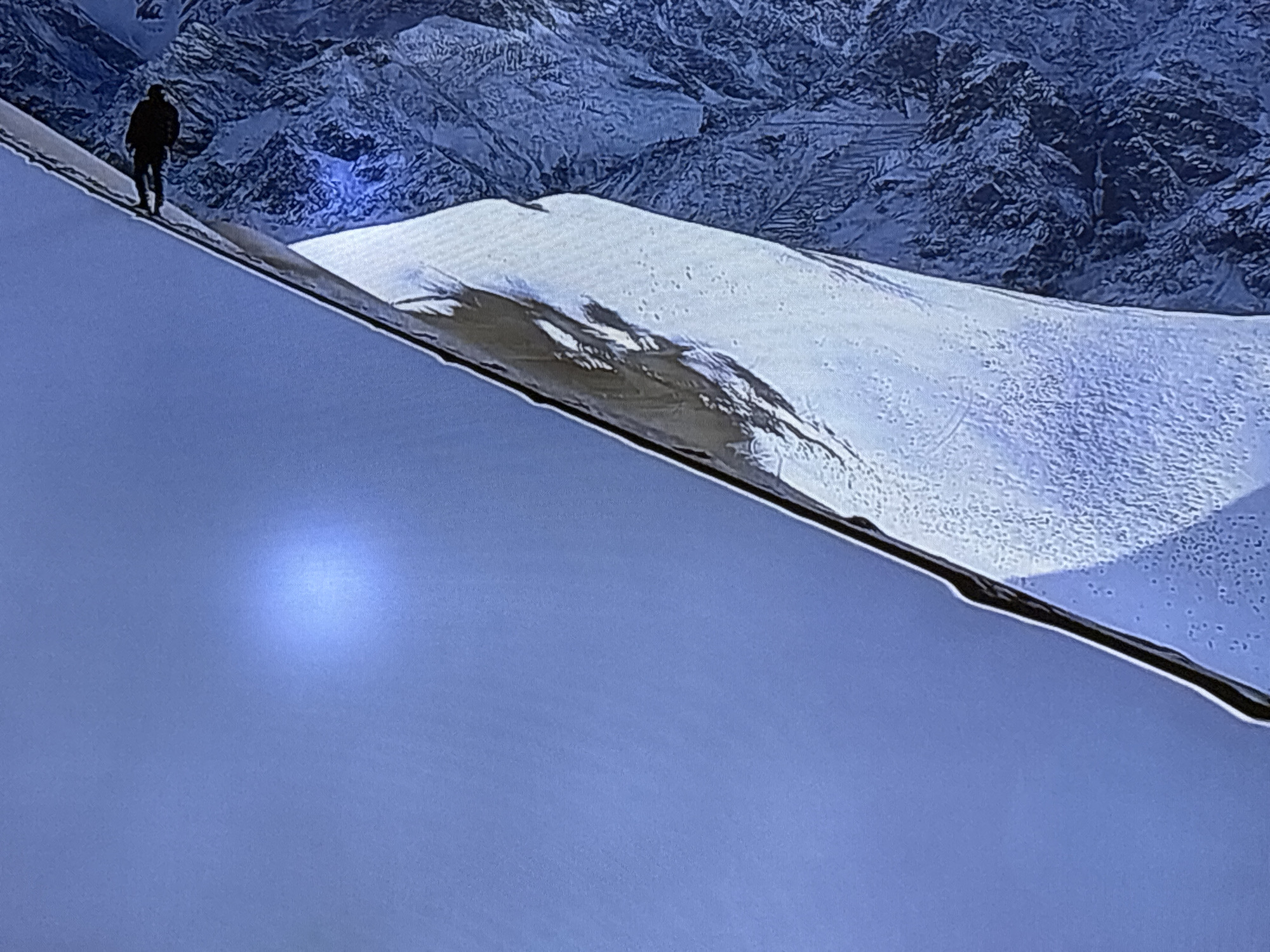 The image shows a lone silhouette of a person standing on a snow-covered mountain ridge. The person is facing away from the camera and appears to be looking out at the vast, snow-covered mountainous landscape stretching into the distance. The mountains in the background are rugged and appear to be covered in snow and ice, with deep shadows indicating the dramatic terrain. The sky is a muted blue, suggesting either early morning or late afternoon light, or possibly overcast conditions. The foreground is dominated by a steep, snow-covered slope that recedes into the distance, creating a sense of depth and isolation. The overall impression is one of a remote and challenging natural environment, possibly a scene from an expedition or a solitary moment of contemplation in a harsh, alpine setting. The image does not contain any elements that would suggest a connection to the city of Kyzylorda, Kazakhstan.