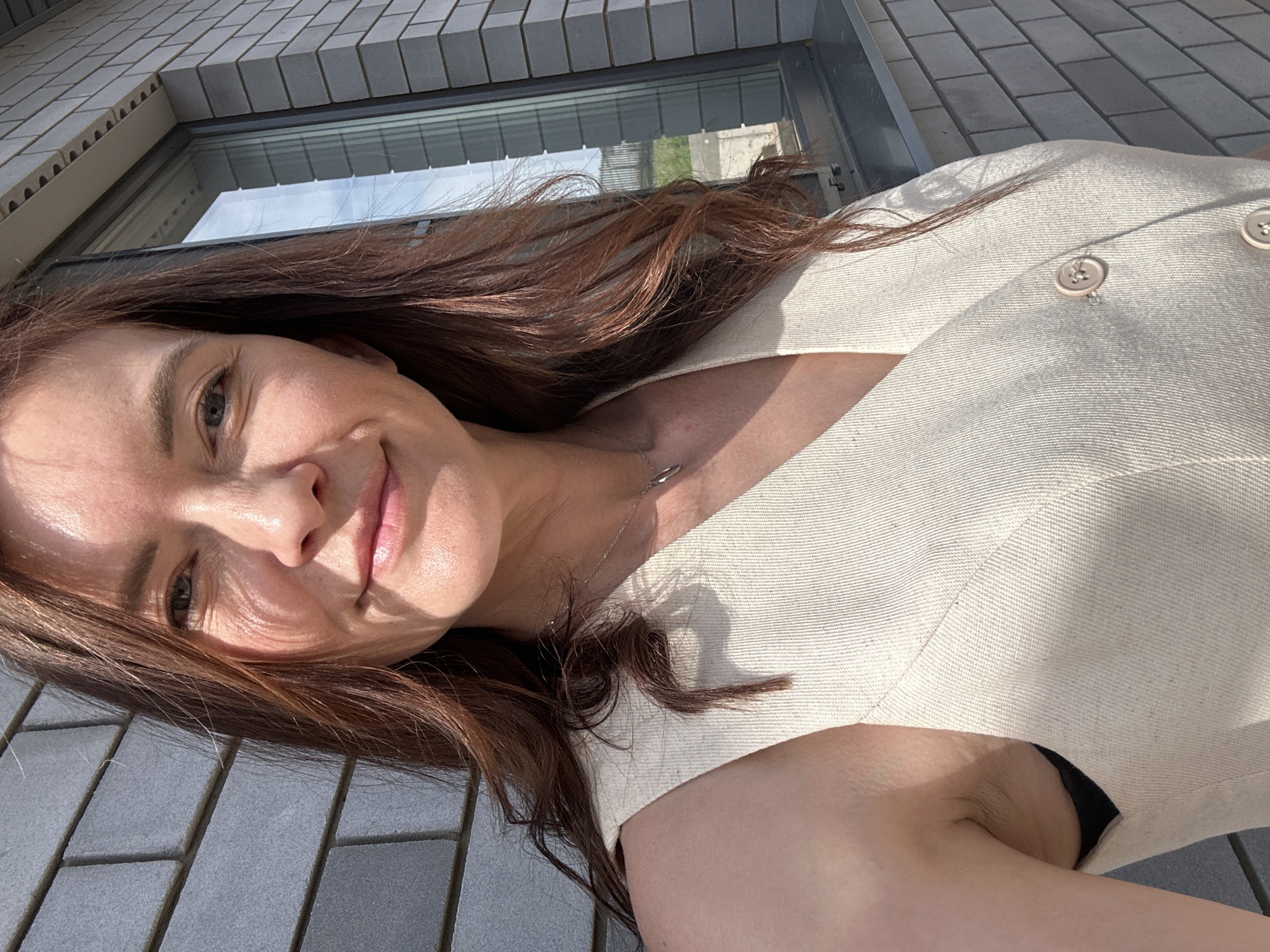 The image depicts a woman in a light-colored, button-down garment, likely a blazer or blouse, posing outdoors. She has long, brown hair and appears to be smiling slightly. The background features a brick-lined building with a window, suggesting an urban environment. The presence of interlocking pavement stones under the sun further points to an outdoor setting. There is nothing in the picture to imply a precise location in the city of Прикамский, Russia.