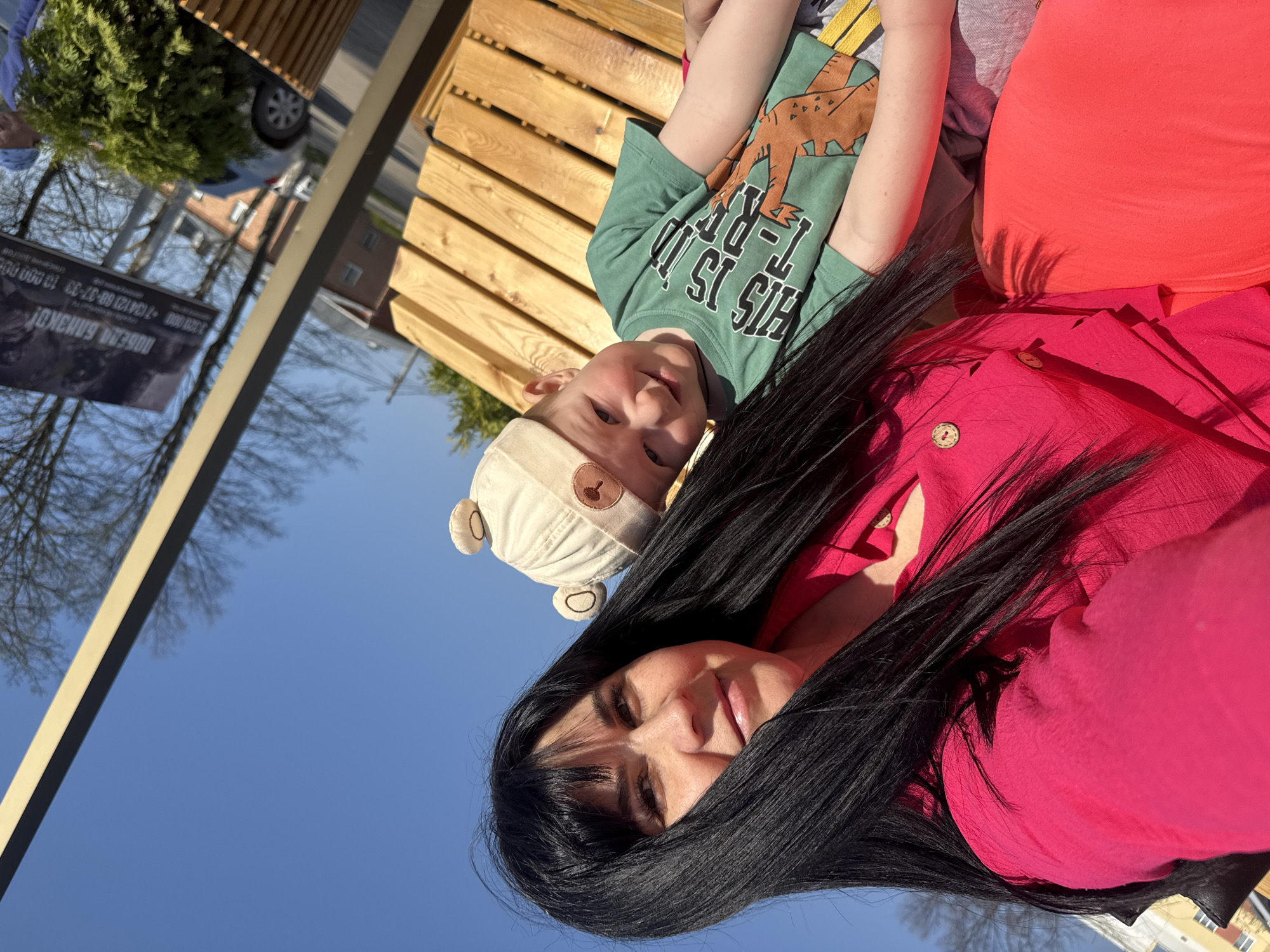 A mother and child pose in the sunshine, with the city of Zav'yalovo, Russia visible in the background. The mother has long black hair and is wearing a magenta-colored blouse with two visible buttons. Her face is prominent, with her dark hair fanning out across her shoulder. The child is next to her, wearing a green T-shirt featuring a brown dinosaur graphic and the words "THIS IS T-REX." He has a white hat with bear ears. The background features a blue sky, trees without leaves, and a wooden structure. A banner is seen in the background with Russian writing on it.