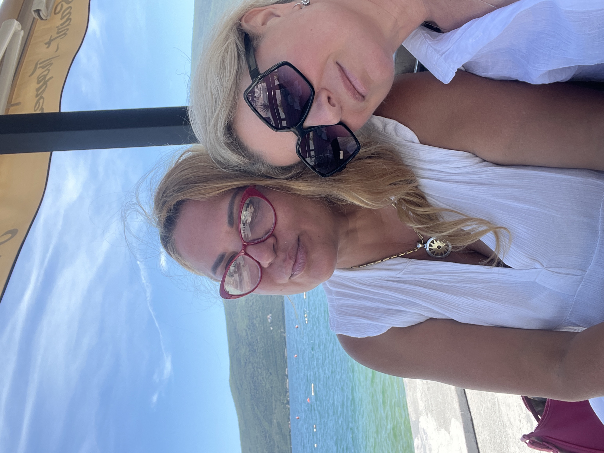 Two Caucasian women are posing for a selfie. One woman has blonde hair, is wearing black sunglasses, and is leaning on the other. The other woman has blonde hair, is wearing red glasses, and a white shirt with a gold necklace. They are outside on what appears to be a pier near the city of Baošići, Montenegro. The sky is blue, and the water is green with a small island in the background. The pier is concrete.