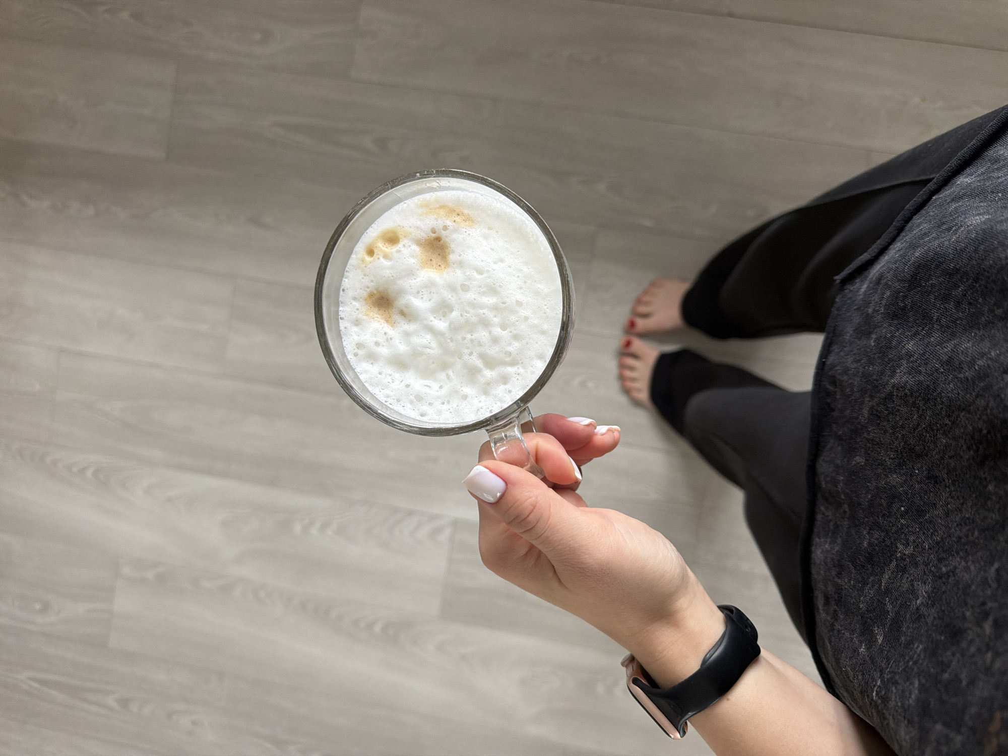 An overhead shot shows a person's hand holding a glass mug filled with a frothy beverage, possibly a latte or cappuccino, with some light brown foam art. The person is wearing a watch on their left wrist and has neatly manicured nails. They are also wearing dark pants and are barefoot, with red nail polish on their toes. The background is a light-colored wooden floor. The image was captured at a residential location in Dreiliņi, Latvia.