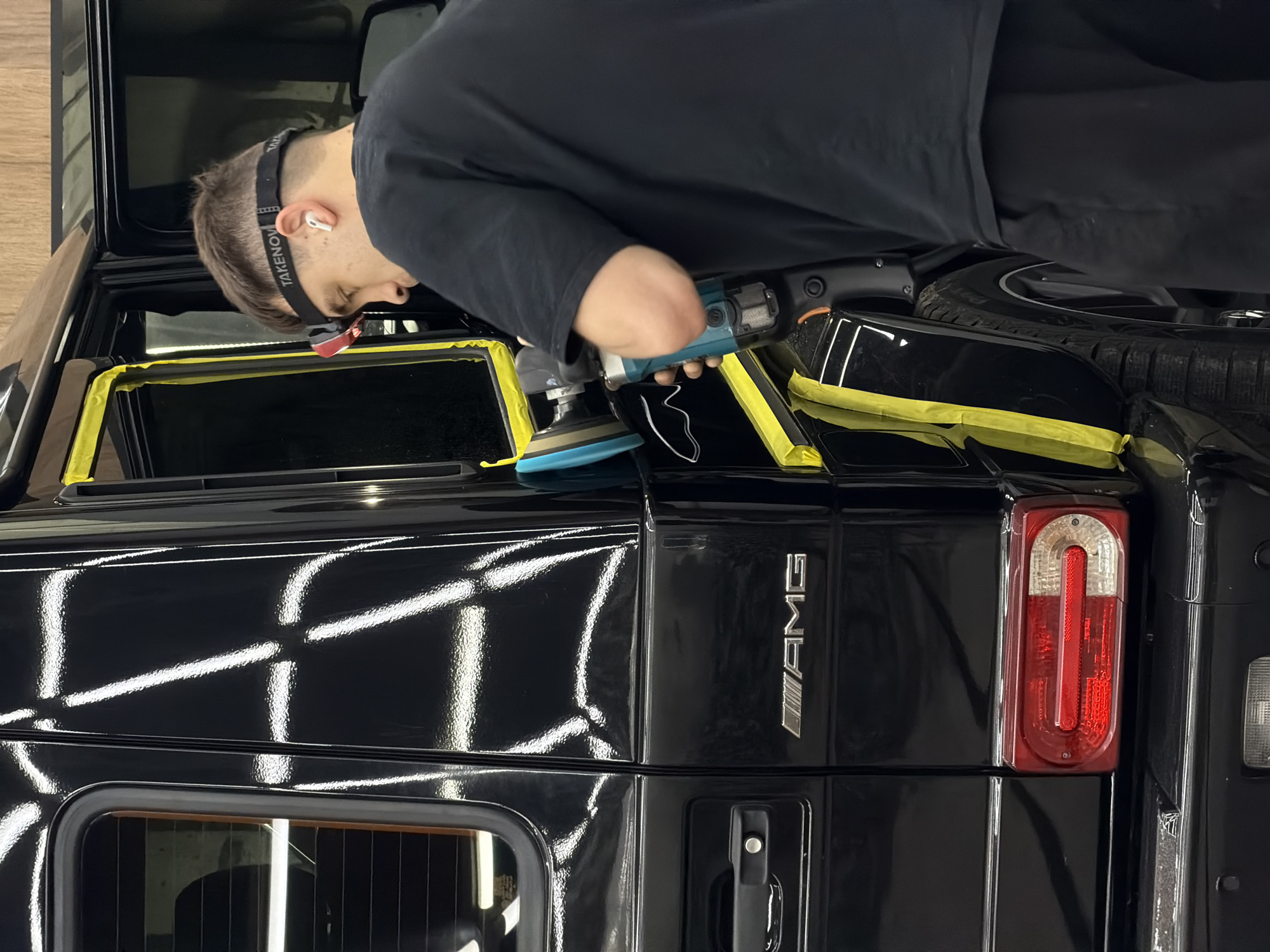 A man is polishing the back of a black Mercedes-Benz G-Class AMG with a blue rotary polisher. Yellow tape is used to protect the edges of the windows. The man is wearing a black long-sleeved shirt and a headlamp. The scene appears to be in a garage or detailing shop. No evidence indicates that the image was taken in the city of Krasnoyarsk, Russia.