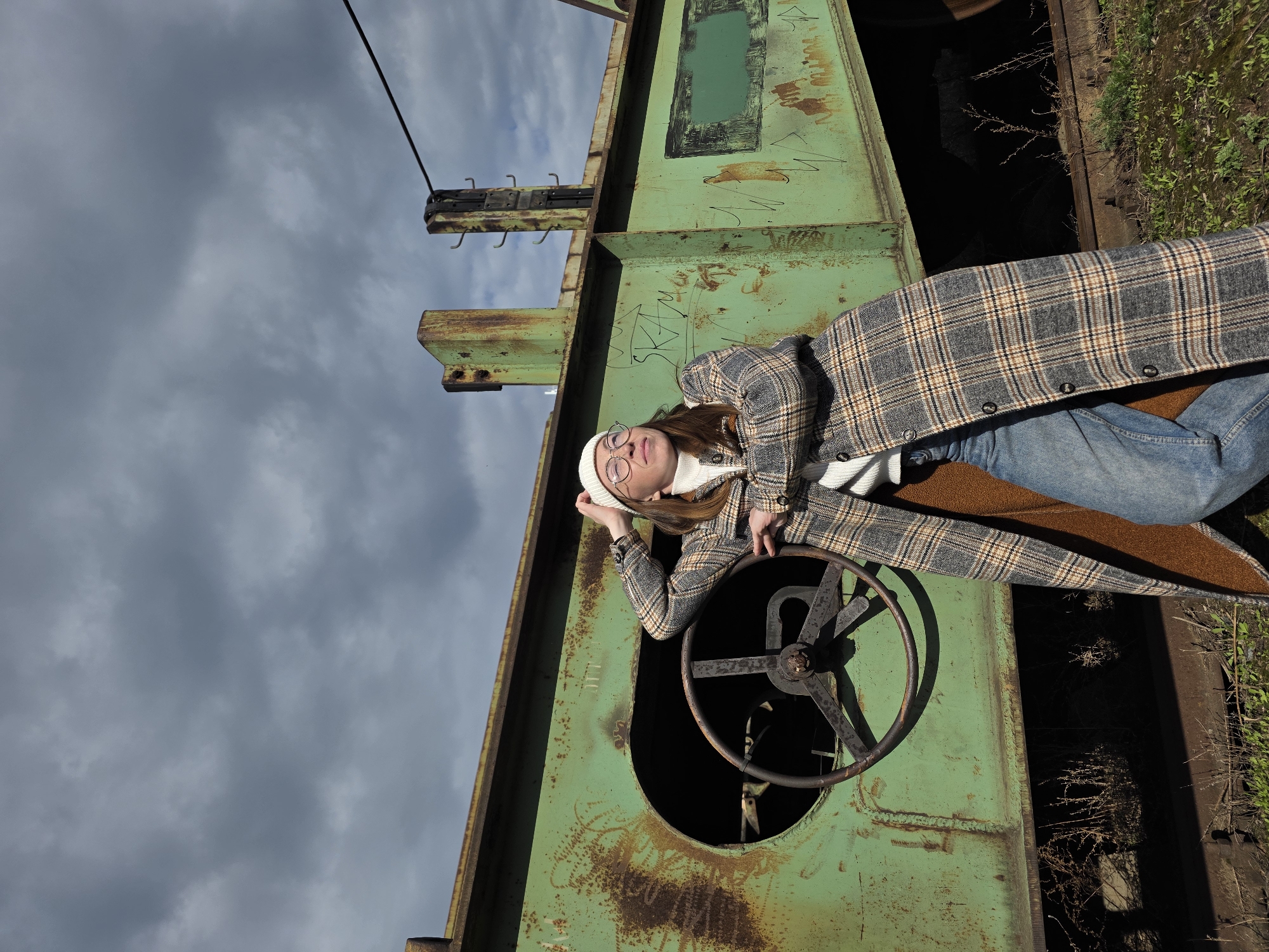 A young woman poses on a large, heavily rusted green metal structure under an overcast sky in what appears to be an industrial or abandoned setting in Chaikovsky, Russia. The woman has light brown hair and wears a white knit beanie, round eyeglasses, a white high-collared top, a plaid long coat in shades of grey, brown, black, and white, and blue jeans. She is lying on her left side on the horizontal section of the structure, with her head slightly tilted back and her gaze directed upwards towards the sky, her left hand near her head in a relaxed pose. Her right arm rests near a prominent, large, dark, spoked wheel integrated into a circular opening on the metal surface.
The robust metal structure, likely a heavy-duty beam or gantry, is painted green but is significantly weathered, showing extensive rust and peeling paint. The sky above is entirely covered by a uniform layer of grey clouds, indicating an overcast day. To the right of the metal structure, a dark, deep void or trench is visible, with sparse, dry vegetation and weeds growing alongside it. Small, rusty metal hooks are also attached to parts of the structure. Several indistinct graffiti markings and faded paint patches adorn the green metal, with letters resembling "SKR" visible on the surface. The scene conveys a contemplative or serene mood, contrasting the stylish attire of the subject with the industrial decay of the environment.