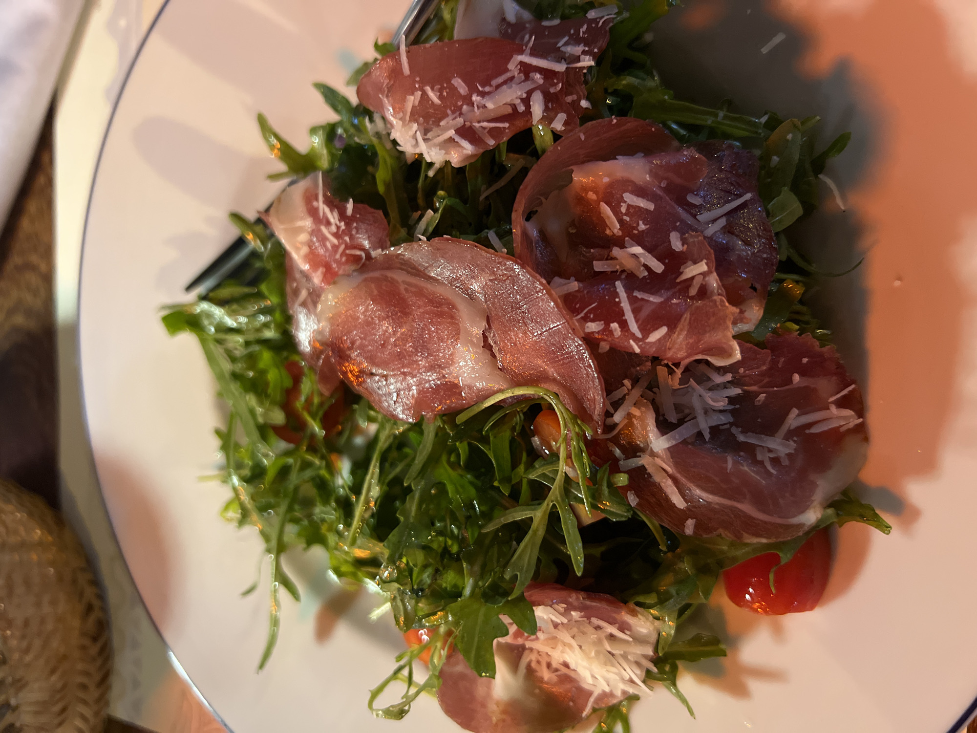 A close-up shot of a white plate featuring a salad. The salad consists primarily of arugula, interspersed with slices of cherry tomatoes and folded prosciutto. Grated parmesan cheese is scattered on top. A fork is visible on the left side of the plate. The scene is illuminated by warm, artificial light. Location information provided suggests this may be occurring in Bac Nha Trang Ward, Vietnam.