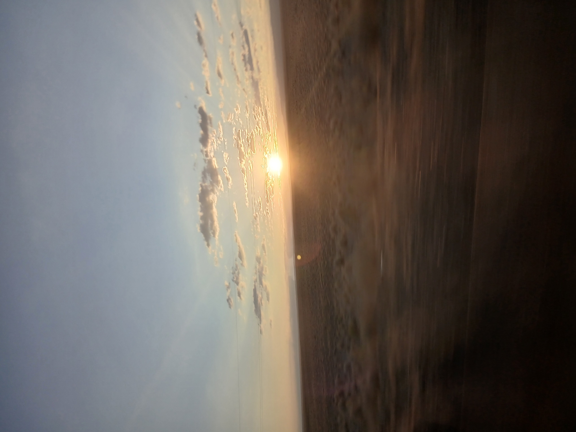 The image captures a sunset over a flat, arid landscape, possibly the steppes surrounding Uzynbulak, Kazakhstan. The sun, a bright, diffused orb, is positioned low on the horizon, casting a warm glow across the sky and illuminating scattered clouds. The sky transitions from a pale blue at the top to a soft yellow and orange near the horizon. The foreground is a blur of brown and tan, suggesting movement, likely from a vehicle. In the midground, low-lying vegetation or dry grasses cover the terrain, appearing slightly blurred due to motion. A faint horizontal line, possibly a power line or a distant ridge, is visible across the landscape. The overall impression is one of a vast, open space at dusk.