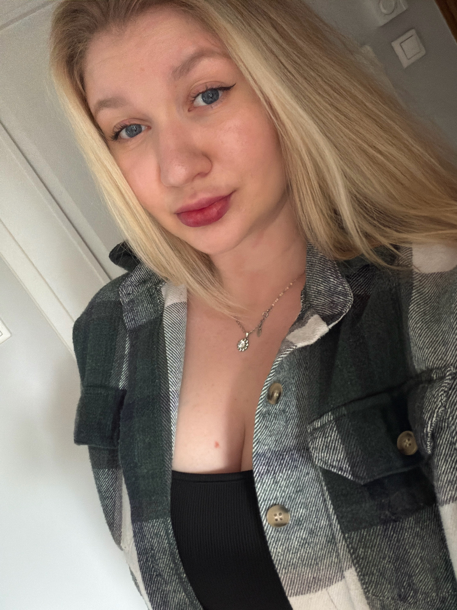 A young woman with blonde hair and blue eyes is captured in a close-up selfie. She is wearing a dark green and white plaid shirt partially open, revealing a black top and a silver necklace with a small pendant. Her lips are painted with dark red lipstick, and she has winged eyeliner. The background appears to be an indoor setting with a white door or wall. The image does not provide any specific visual cues that would allow for its location to be identified within the city of Koszalin, Poland.
