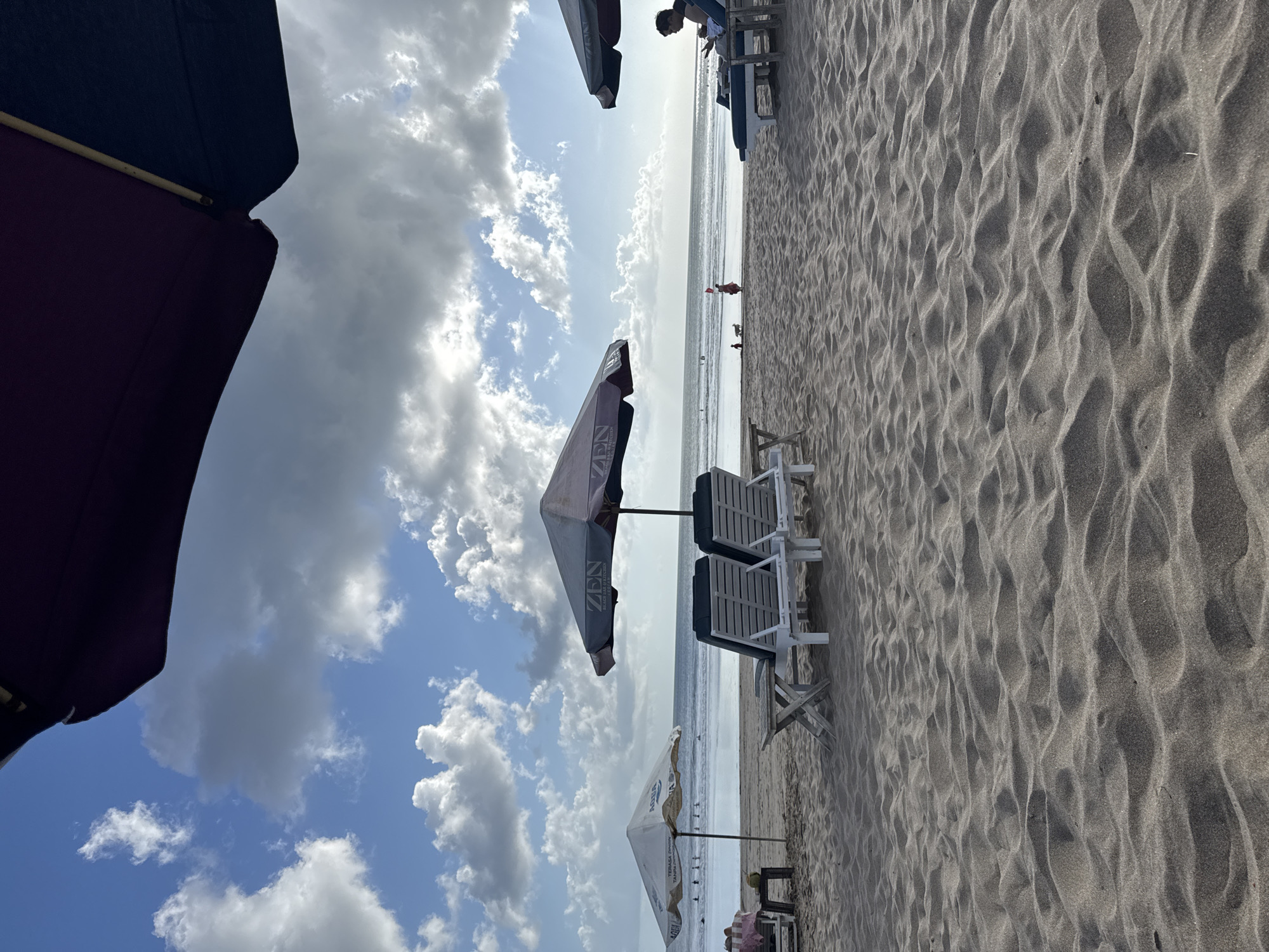 The image depicts a beach scene in Kuta, Indonesia, with a focus on the sandy shore, umbrellas, and lounge chairs. The sky is partly cloudy with a mix of white and grey clouds against a blue backdrop. In the foreground, rippled sand dominates the lower portion of the frame.
Several beach umbrellas are visible. One large, dark umbrella extends from the top right corner of the image, obscuring a portion of the sky. Closer to the middle ground, a large umbrella with "ZEN" printed on it shades two white lounge chairs. To the left, a smaller white umbrella, possibly branded "AQUA," shades a single lounge chair.
There are a few people present in the scene. On the far right, a person is seated on a lounge chair, appearing to be looking at a phone. In the mid-ground, near the water's edge, a few small figures can be seen, including what appears to be a child and a dog. Further out in the water, several individuals are visible, suggesting swimming or surfing activities. The ocean stretches to the horizon, with gentle waves lapping the shore. The overall impression is a relaxed beach atmosphere.