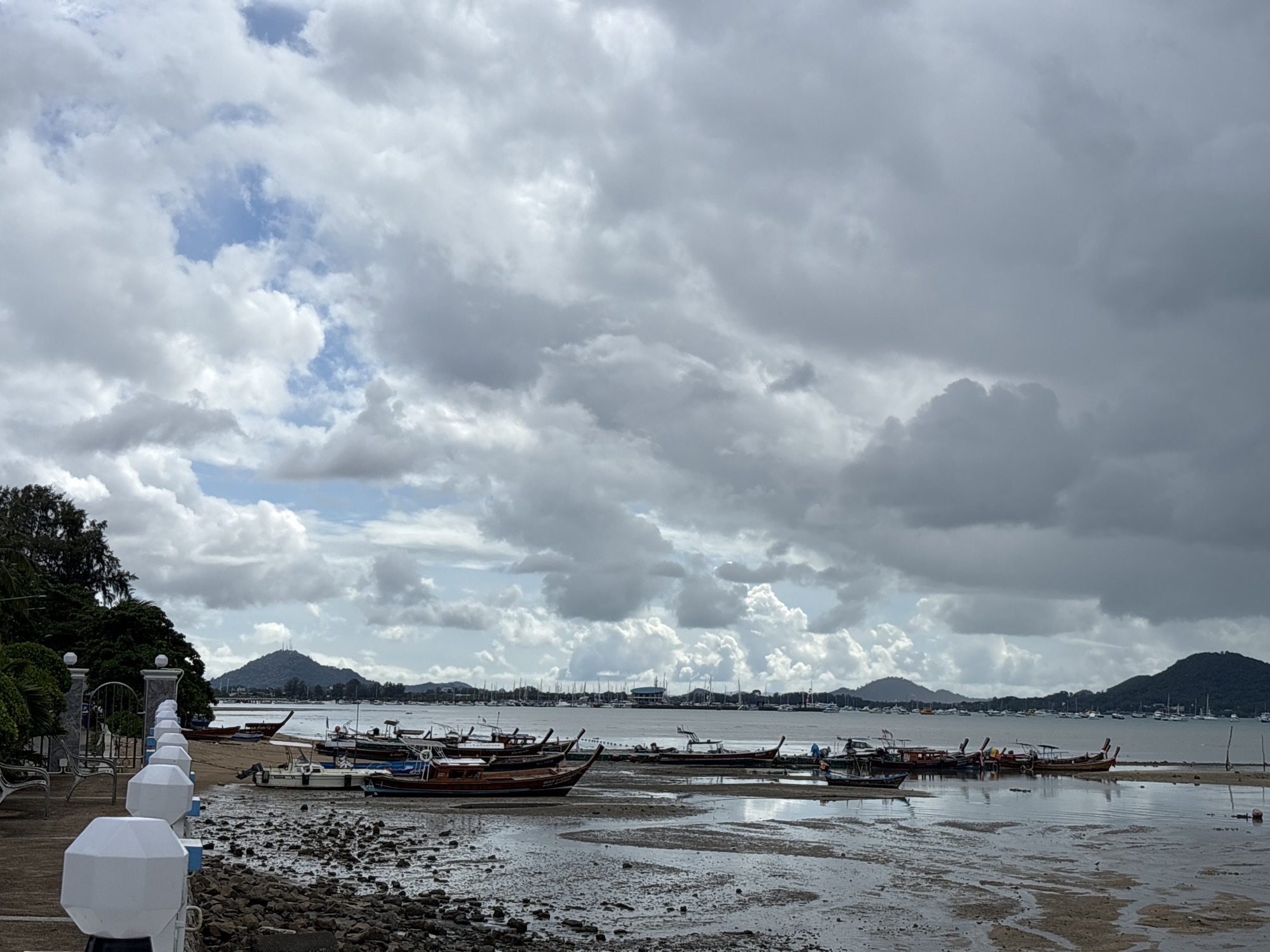 This is an overcast, medium-shot photograph of a muddy shoreline in Rawai, Thailand, with numerous longtail boats grounded and several smaller boats anchored offshore. The foreground is dominated by the textured, wet sand and scattered dark rocks. In the middle ground, a collection of wooden longtail boats, varying in size and color from dark brown to lighter wood tones, are pulled up onto the mud. Some of these boats have small motors attached. A few white motorboats are also visible among them. In the background, a calm body of water stretches towards a distant shoreline lined with lush green hills. A larger group of sailboats and other vessels are anchored further out. The sky is filled with a dramatic display of heavy, grey clouds, with patches of brighter, lighter clouds and hints of blue sky peeking through. On the left side of the image, a paved walkway with white decorative posts and a metal fence runs along the edge of the shore, with benches placed near the fence. Green trees and foliage are visible behind the walkway. The overall scene suggests a quiet coastal area, possibly at low tide, under a cloudy sky. No people are clearly visible in the image.