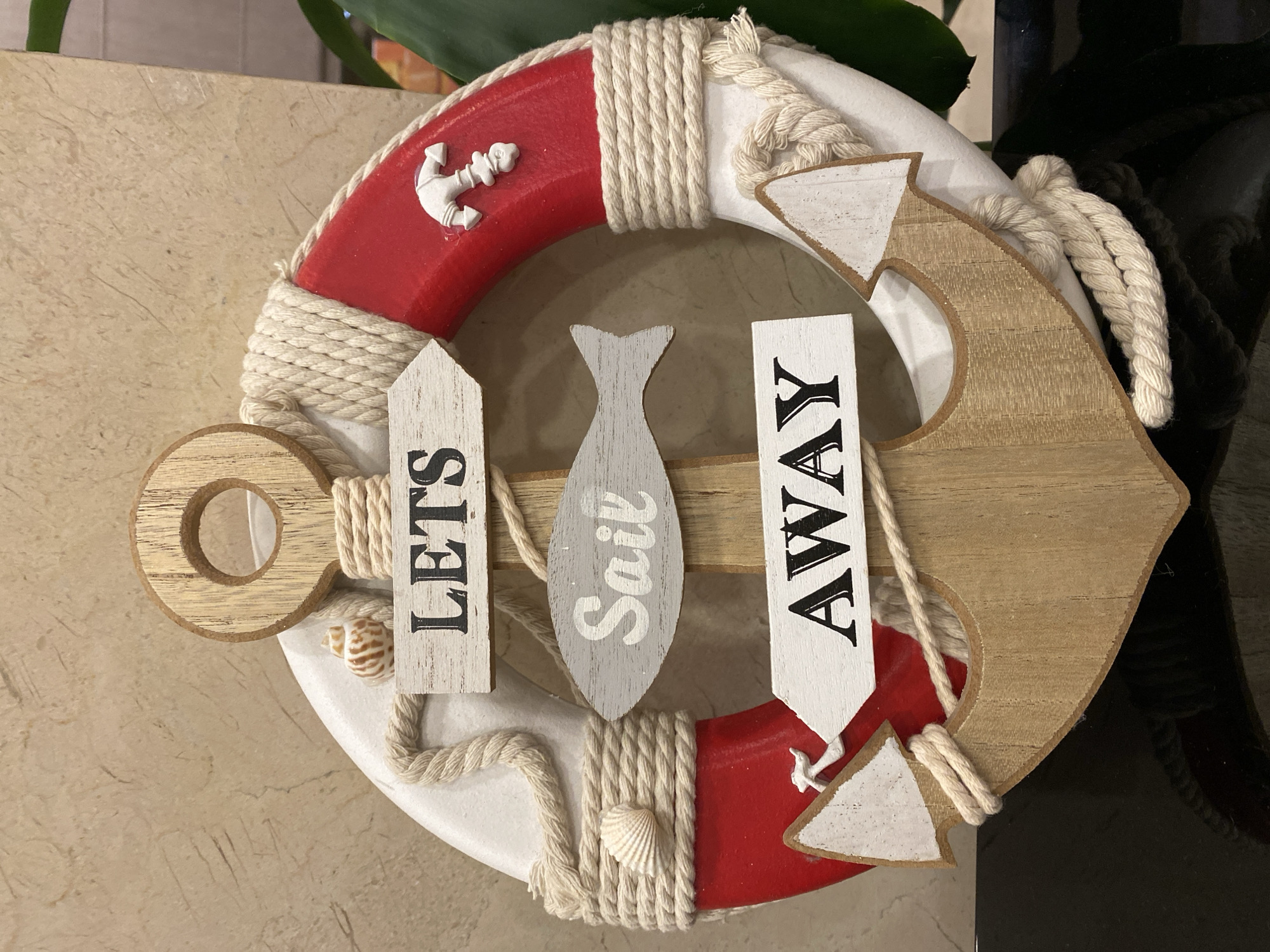 A nautical-themed decorative object, likely for a home or business, is displayed against a textured, beige wall. The object is composed of a red and white life preserver, wrapped with thick, cream-colored rope. A wooden anchor shape is attached to the bottom of the life preserver. Attached to the front of the object are three wooden signs. The top sign, pointing left, reads "LETS" in black block letters. The middle sign, shaped like a fish and colored gray, reads "Sail" in white cursive letters. The bottom sign, pointing left, reads "AWAY" in black block letters. A small white anchor figurine is attached to the red portion of the life preserver. Small seashells are also incorporated into the decoration, one near the "LETS" sign and another on the rope to the left of the "Sail" sign. The scene does not provide enough information to determine a specific location within the city of Đà Nẵng, Vietnam.