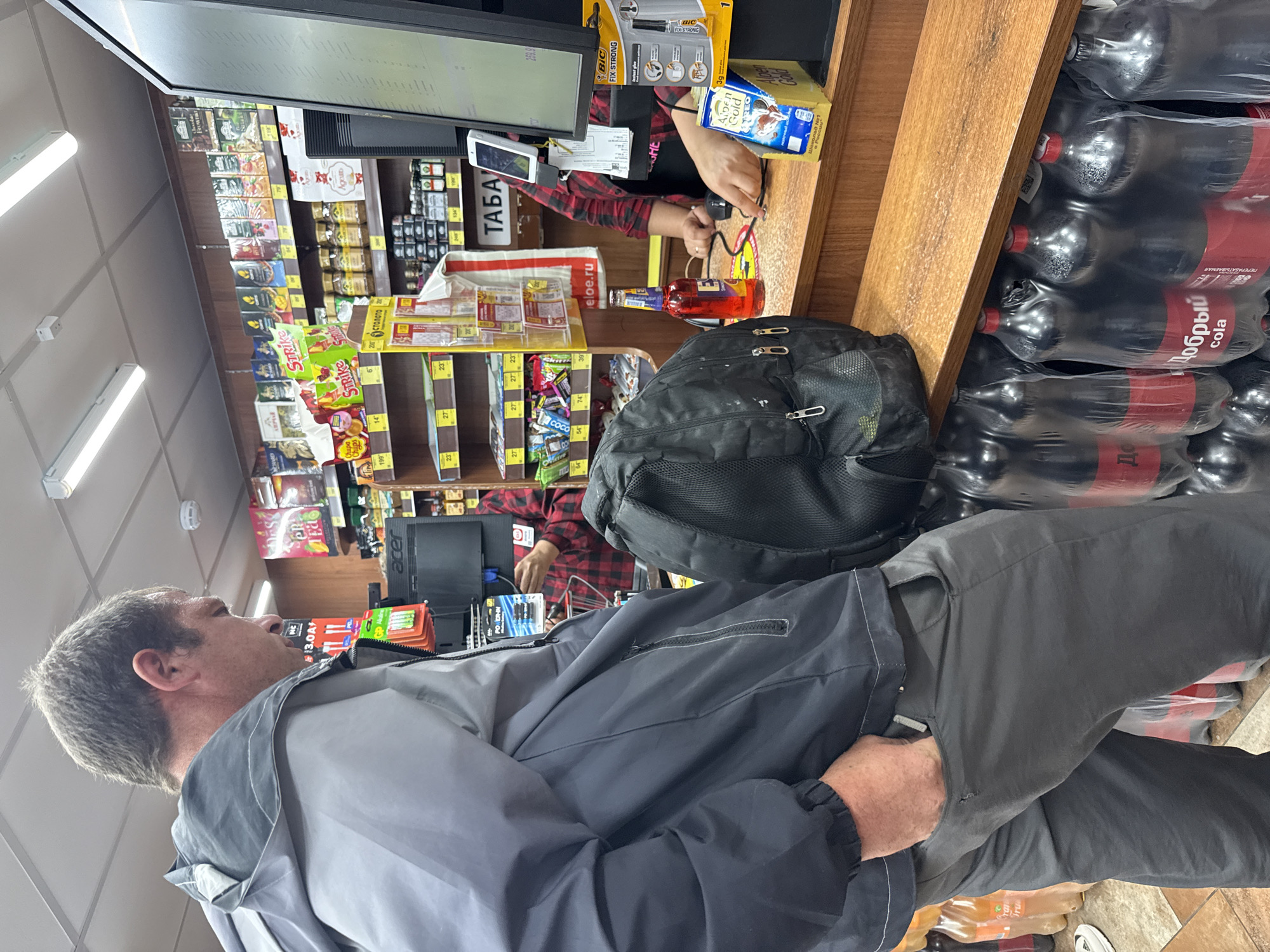 A man wearing a grey and black jacket and grey pants stands at a checkout counter in what appears to be a convenience store. He has his left hand in his pants pocket and his right hand is not visible. He is facing away from the camera, looking towards the checkout counter. Behind him, a backpack is slung over his shoulders. A cashier, wearing a red and black plaid shirt, is behind the counter, interacting with the man. There are shelves filled with various products behind the cashier. In the foreground, there are rows of plastic bottles, likely beverages, stacked on shelves. The environment appears to be indoors, well-lit with fluorescent lighting. The image does not provide any explicit details to confirm the location as Moscow, Russia, but the signage and product packaging might suggest it.