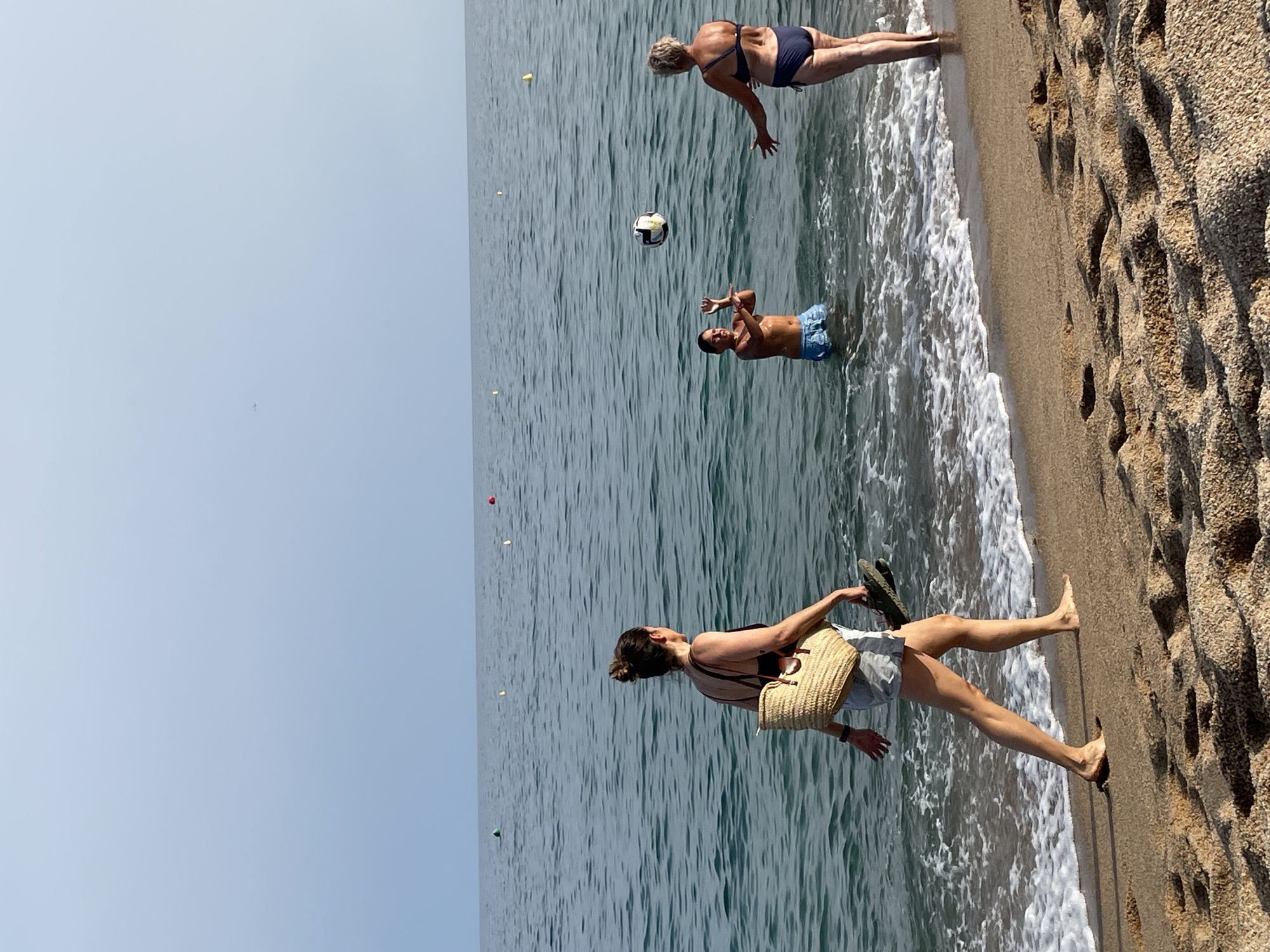 In Canet de Mar, Spain, on a clear, sunny day, three people are enjoying the beach and the sea. The scene is a coastal setting with the Mediterranean Sea on the left and a sandy, pebbly beach on the right.
In the water, two individuals are playing volleyball. A woman with blonde hair, wearing a dark blue bikini, is positioned further out in the water, her body angled as if reaching for the ball. Nearby, a man in blue swim trunks is in the water, his arms raised, seemingly playing the game with her. A volleyball is suspended in the air between them.
Closer to the shore, a woman is walking along the water's edge. She is wearing a black tank top and light-colored shorts, carrying a straw tote bag over her shoulder and a pair of dark sandals in her right hand. Her legs are bare, and she is stepping into the shallow water, with small waves gently lapping at the shore. Her feet are bare.
The time of day appears to be midday or early afternoon, suggested by the bright sunlight casting minimal shadows. The weather is pleasant, characteristic of a summer day at the beach. The water is a deep blue-green, with small white crests of waves breaking near the shore. The beach itself is composed of sand and small pebbles, with some larger, rounded stones and indentations in the sand. The overall mood is one of leisure and recreation.