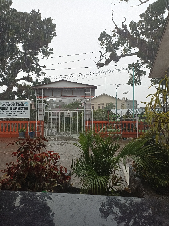A daytime scene depicts the entrance of State Elementary School 11 Banda Sakti (SD Negeri 11 Banda Sakti) at Yahuda Street No. 2, Huta Blang Village, Banda Sakti District, Lhokseumawe City, Indonesia. The weather is rainy, with visible raindrops against an overcast sky.
In the immediate foreground, a dark, wet, reflective surface, possibly stone or concrete, partially obscures green and red-leafed ornamental plants. A small palm-like plant is prominent on the right.
The midground is dominated by a white metal gate with vertical bars, leading into the school grounds. Above the gate, a light-colored, gabled archway structure is present. To the left of the gate, a white rectangular sign with black lettering identifies "PEMERINTAH KOTA LHOKSEUMAWE DINAS PENDIDIKAN DAN KEBUDAYAAN" (Government of Lhokseumawe City, Education and Culture Office) and "SD NEGERI 11 BANDA SAKTI," along with its address and NPSN: 10105805. An orange-painted fence, topped with vertical balusters, extends horizontally from the gate. On the right section of this fence, a vertical garden system with multiple light green racks holds numerous small potted plants. The ground beyond the gate is wet, dark-colored pavement.
The background reveals several buildings, including a light-colored structure with a beige facade and a dark sloped roof on the right. Numerous parallel power lines or utility cables stretch across the upper portion of the frame. Large, densely foliated trees, visibly wet from the rain, frame the scene on both the left and right sides. A green-poled street light with an attached satellite dish is visible behind the fence on the right. No human subjects are present in the image.