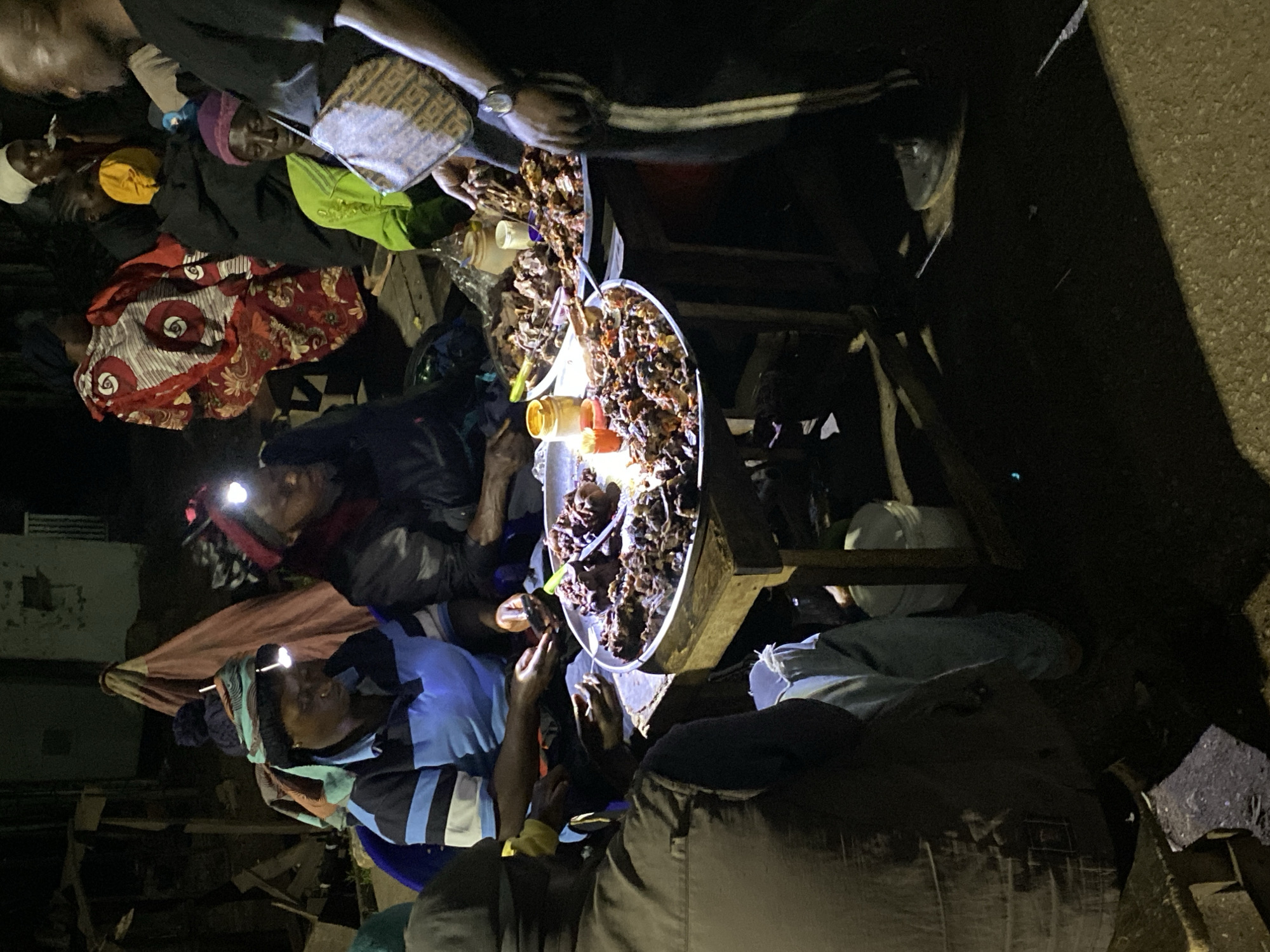 The image shows a nighttime scene in Bukuru, Nigeria. Several people are gathered around a metal food stall supported by wooden legs. Two large trays hold what appears to be cooked meat or similar food items, illuminated by a central light source and headlamps worn by some of the individuals. People are reaching for the food, possibly making purchases. Various containers and personal items are scattered around the base of the stall. The surrounding environment is dark and somewhat cluttered.