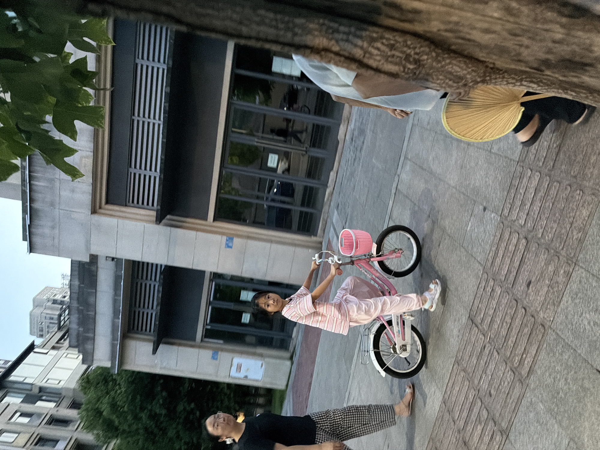 In the city of Jiangjin, China, a young girl rides a pink bicycle with a basket on the handlebars along a paved sidewalk. She wears a striped pink shirt and matching pants. To her left, an adult woman walks, wearing a black t-shirt and checkered pants. In the background, modern buildings with large windows and some greenery are visible. The scene appears to be outdoors, possibly in a park or on a street. Another person is partially visible in the foreground on the right, holding a yellow fan.
